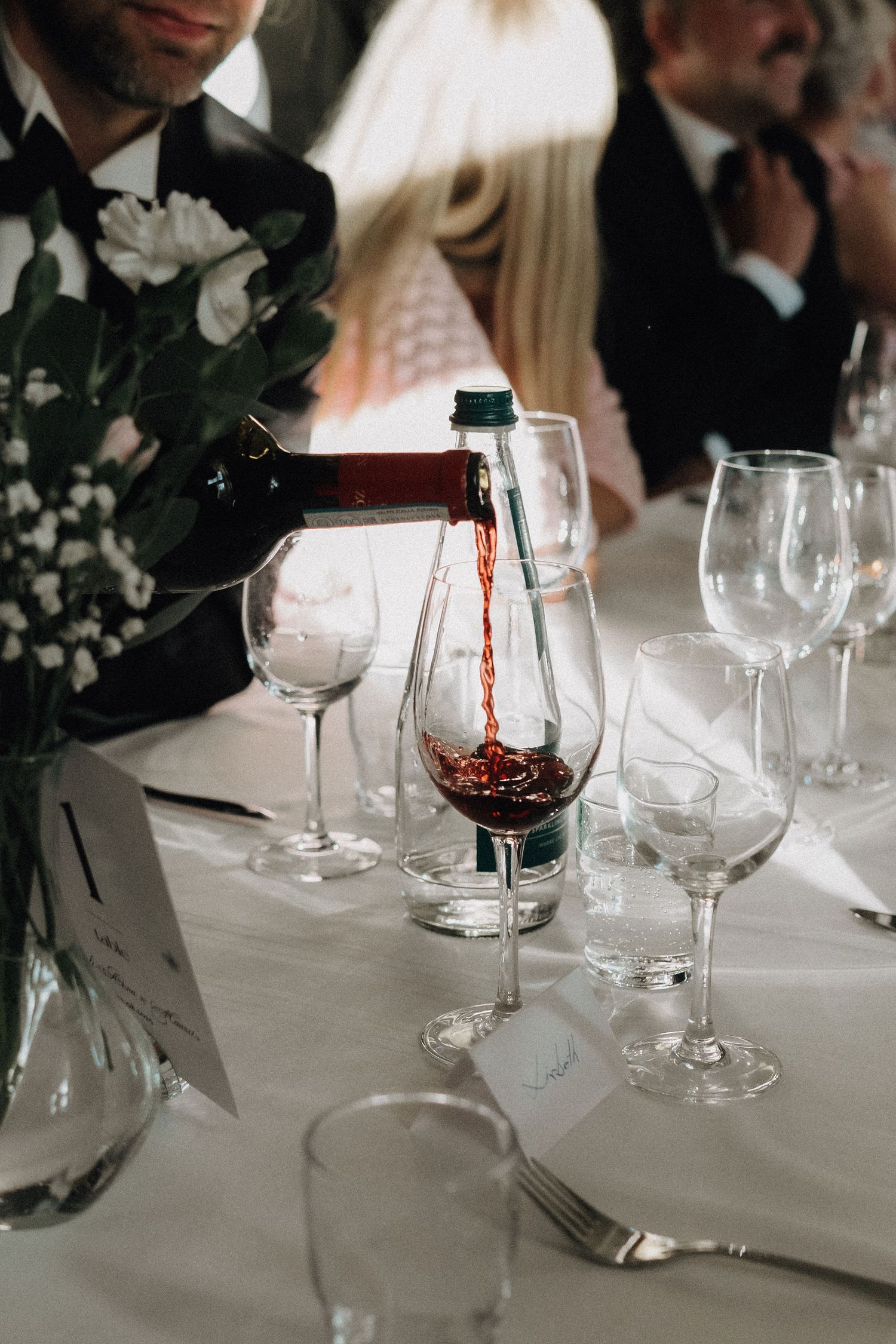 Red wine being poured into a glass at an elegant wedding reception dinner table with crystal glassware.