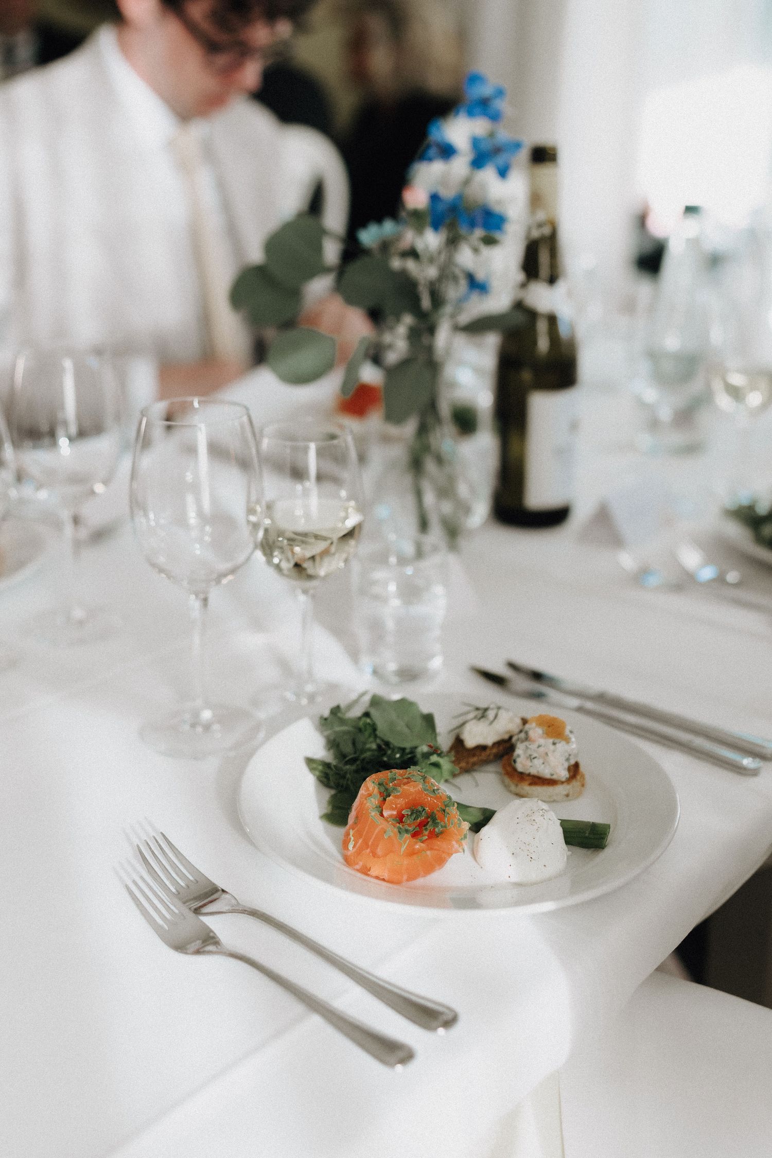 Elegant wedding reception table setting with salmon appetizer on white plate, crystal wine glasses, and floral centerpiece.