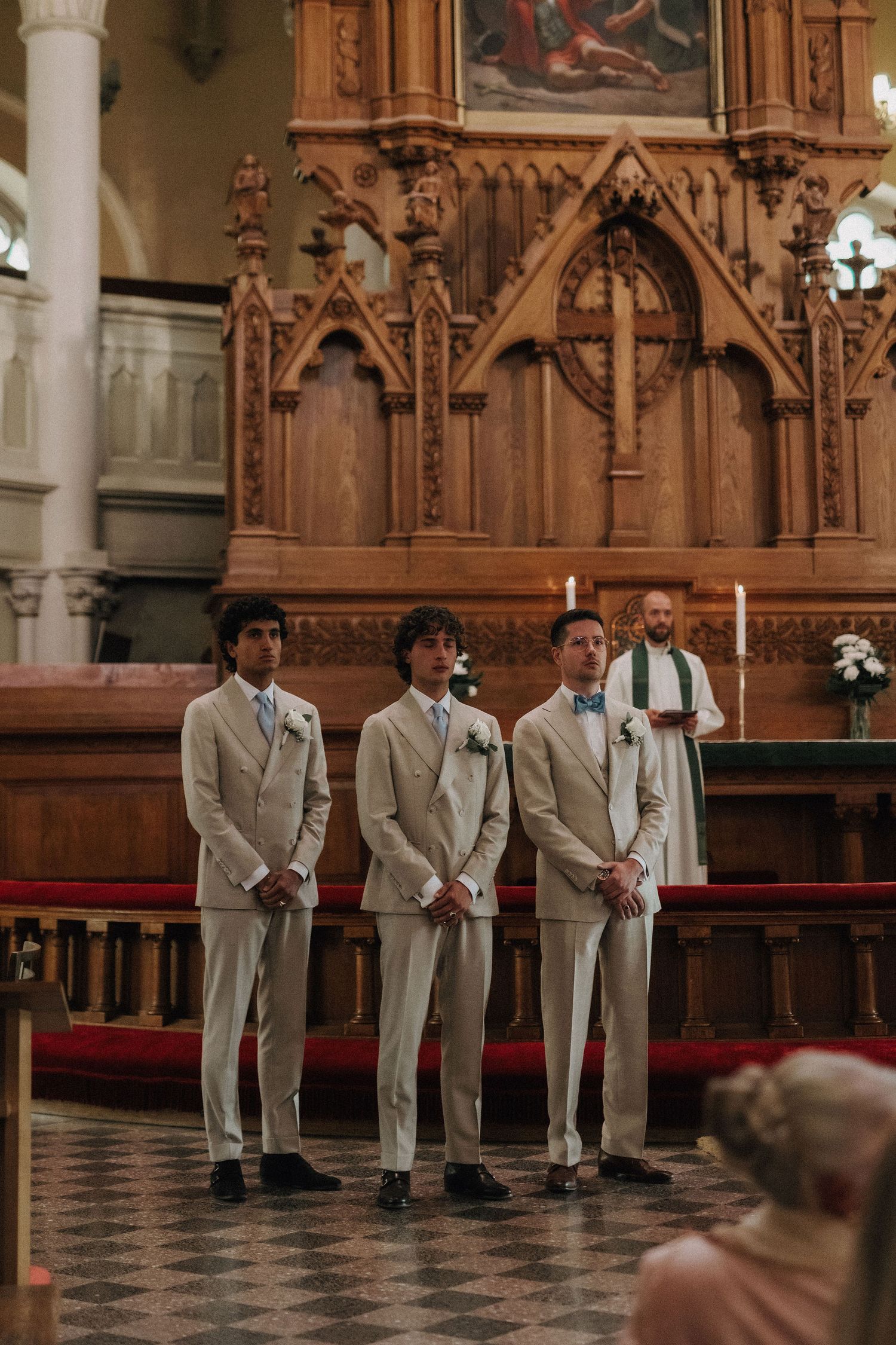 Three groomsmen in cream suits stand at church altar before ornate wooden reredos.