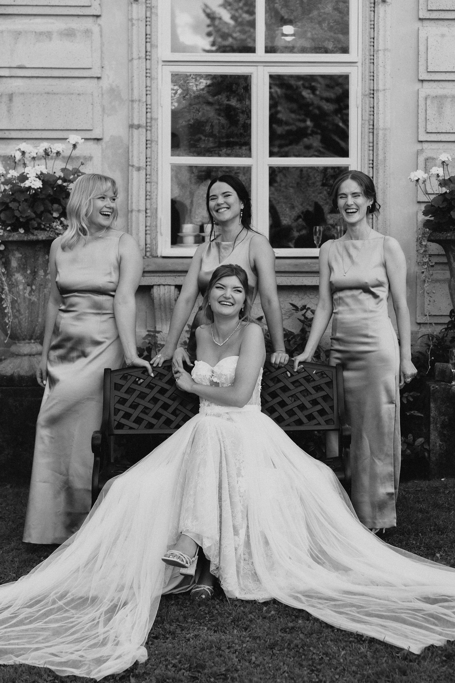 Bride seated on bench laughing with three bridesmaids in satin gowns outside elegant venue, black and white photo.