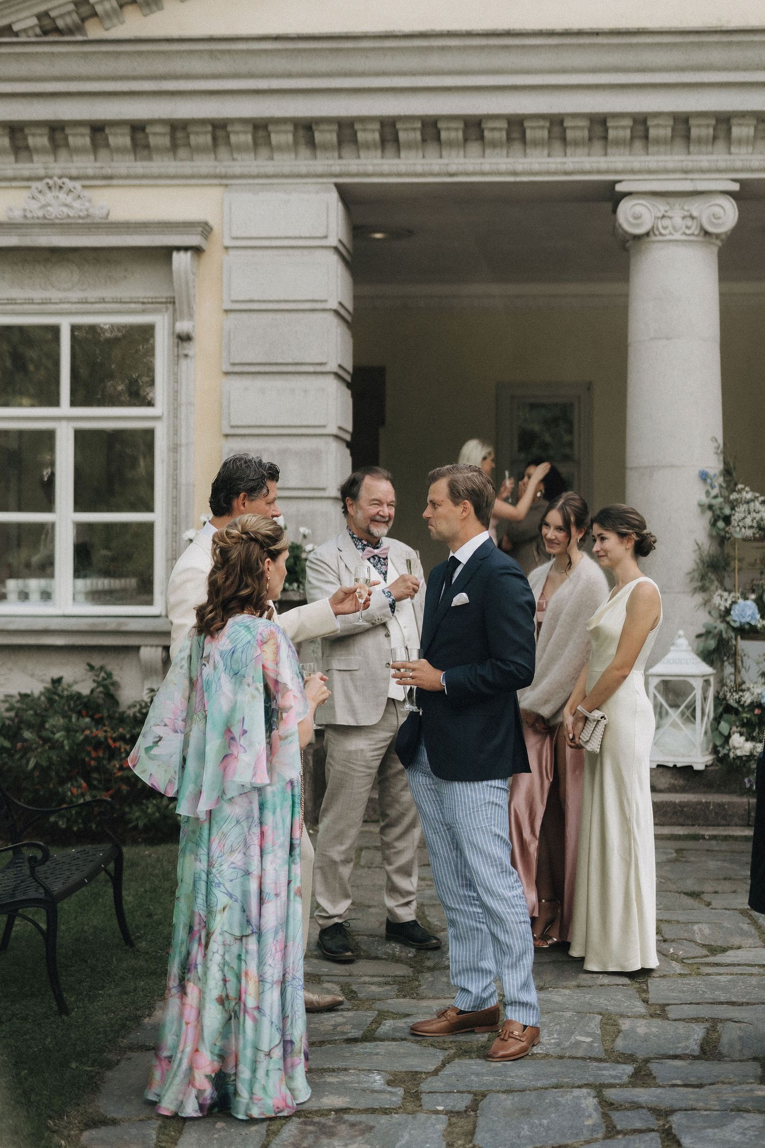 Wedding guests socialize outside a neoclassical venue as a woman in a floral dress converses.
