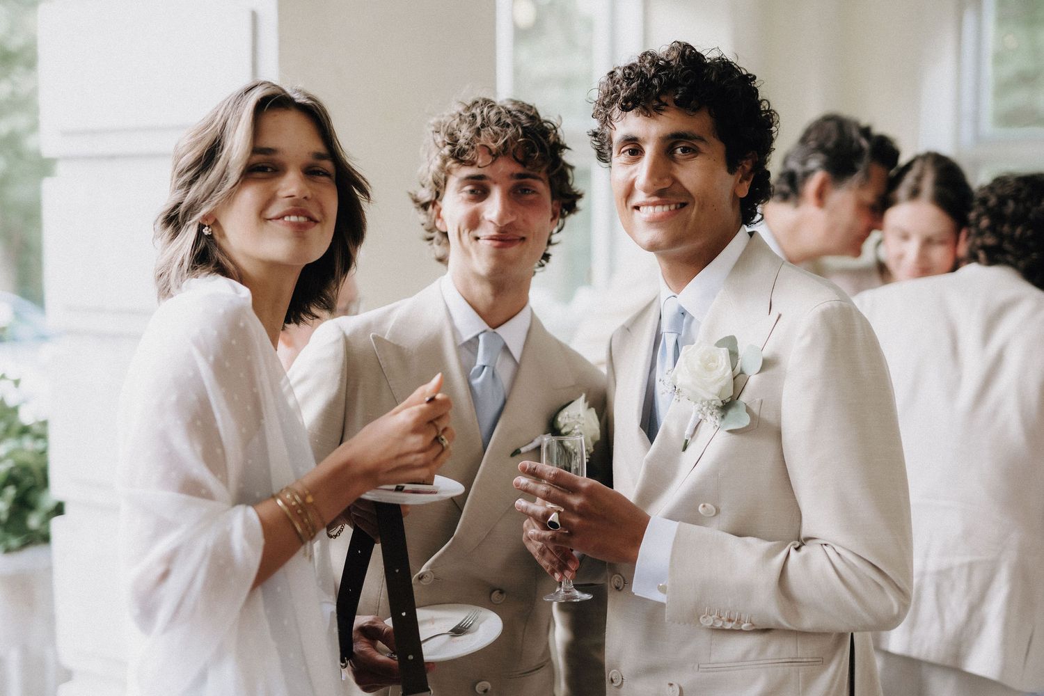 Bride and two groomsmen in beige suits smile together holding champagne glasses at wedding reception.