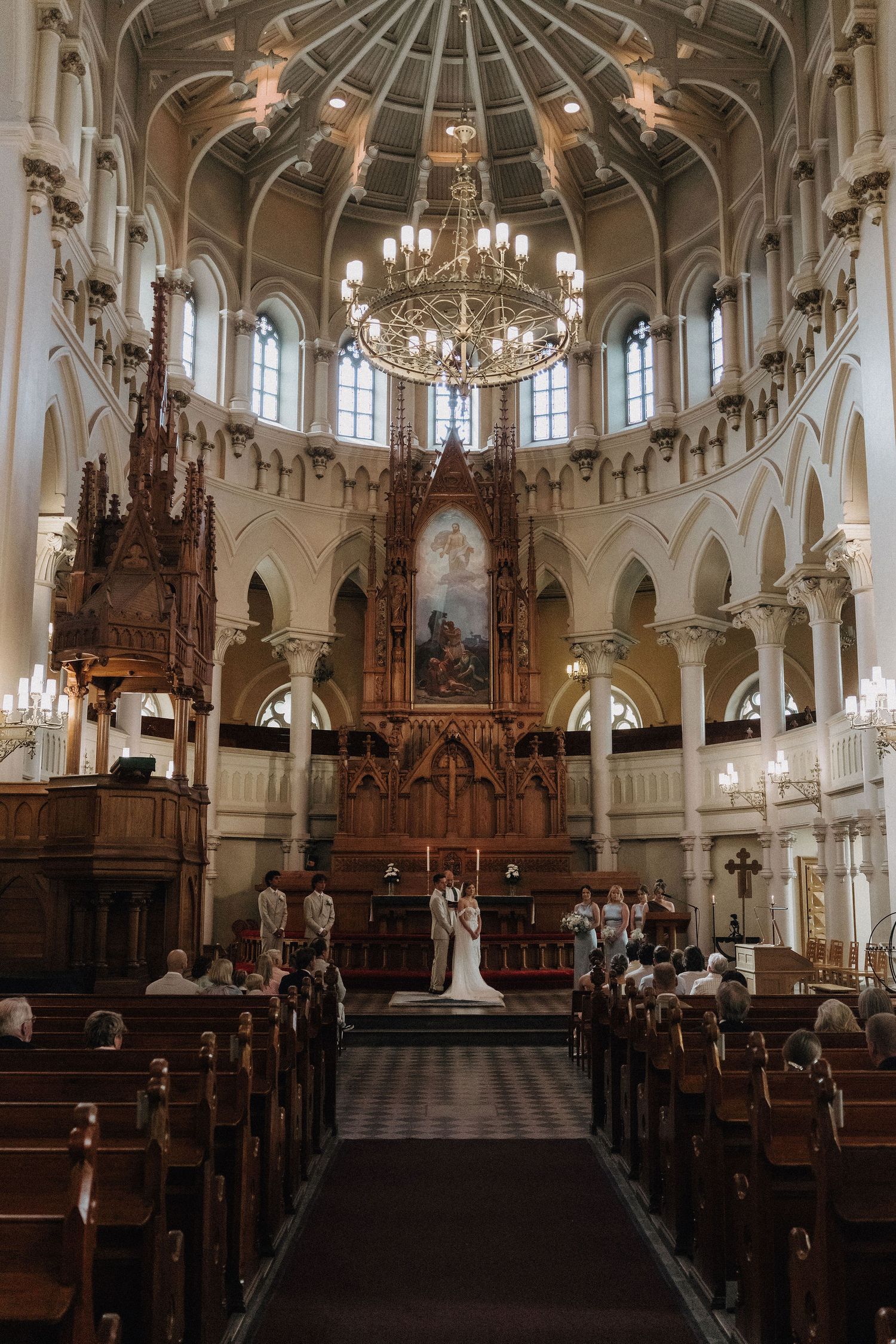 Wide shot of elegant church wedding ceremony under grand chandelier with ornate altar and wooden pews filled with guests.