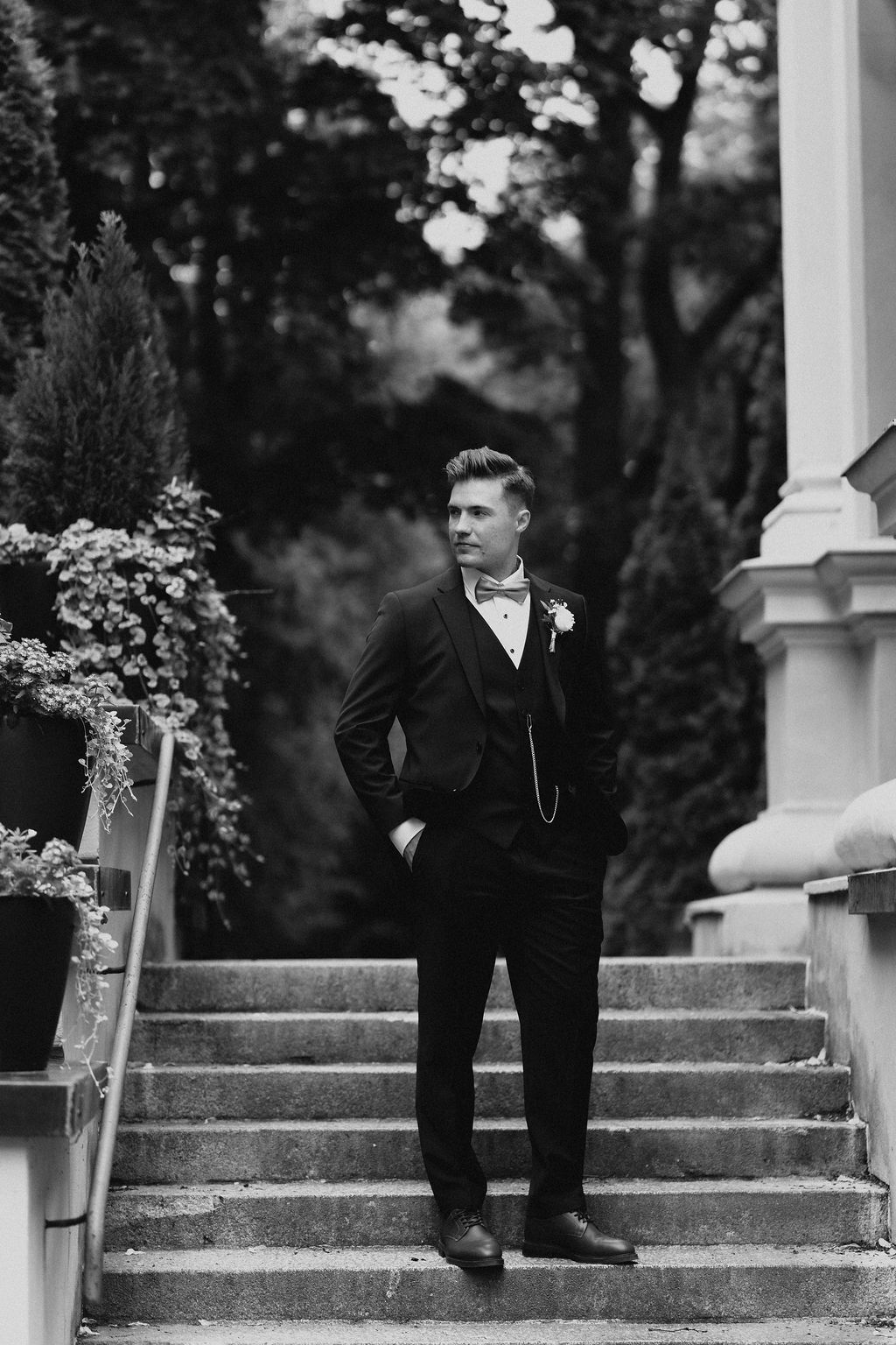 Elegant groom in black suit with bow tie poses on stone steps in black and white portrait.
