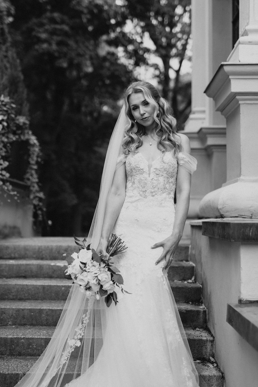 Black and white portrait of blonde bride in lace mermaid gown holding bouquet on stone stairs.