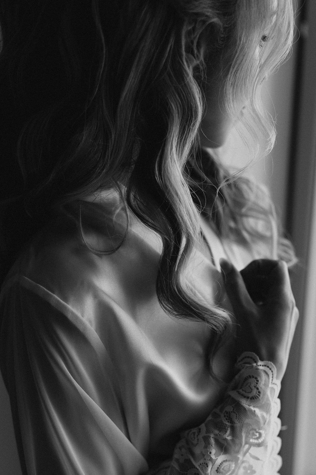 Artistic black and white portrait of bride in lace robe gazing out window before wedding.