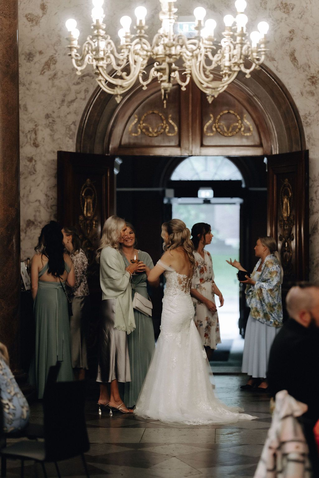 Bride in fitted lace gown socializing with guests under crystal chandelier in elegant venue.