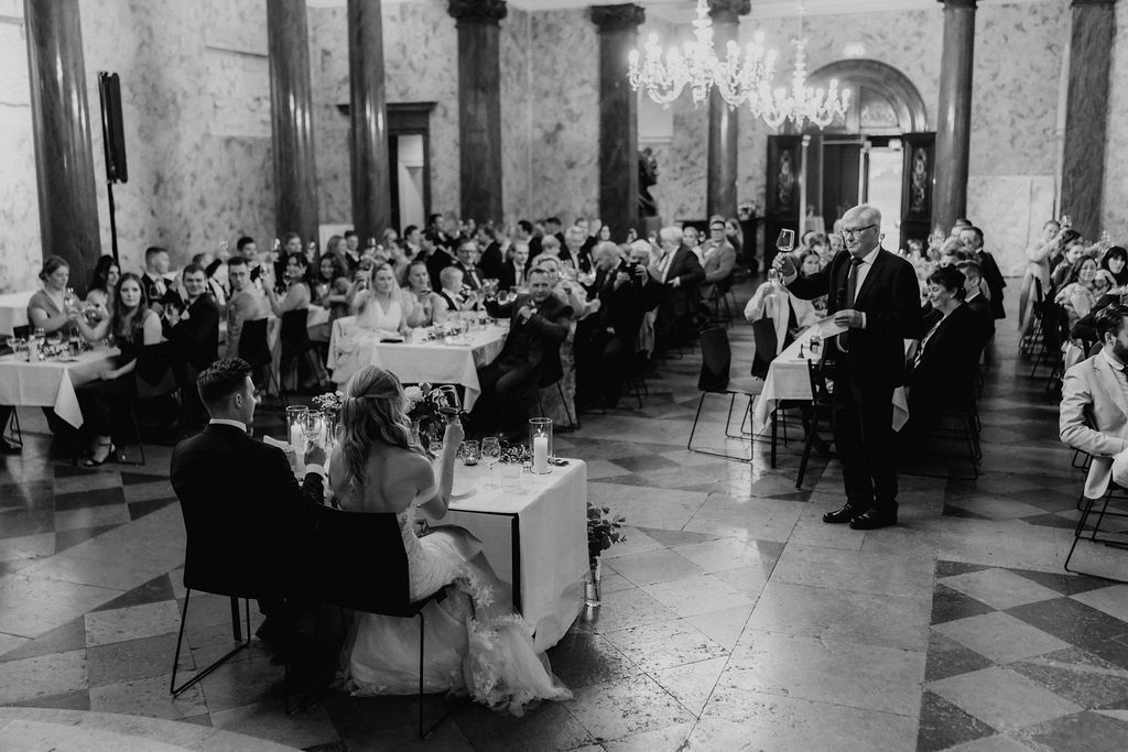 Wide shot of wedding reception in grand marble hall with guests applauding during speeches.