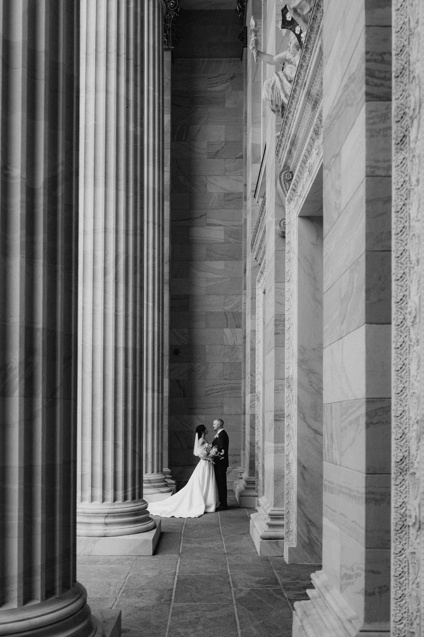 Black and white photo of bride and groom embracing between grand stone columns.
