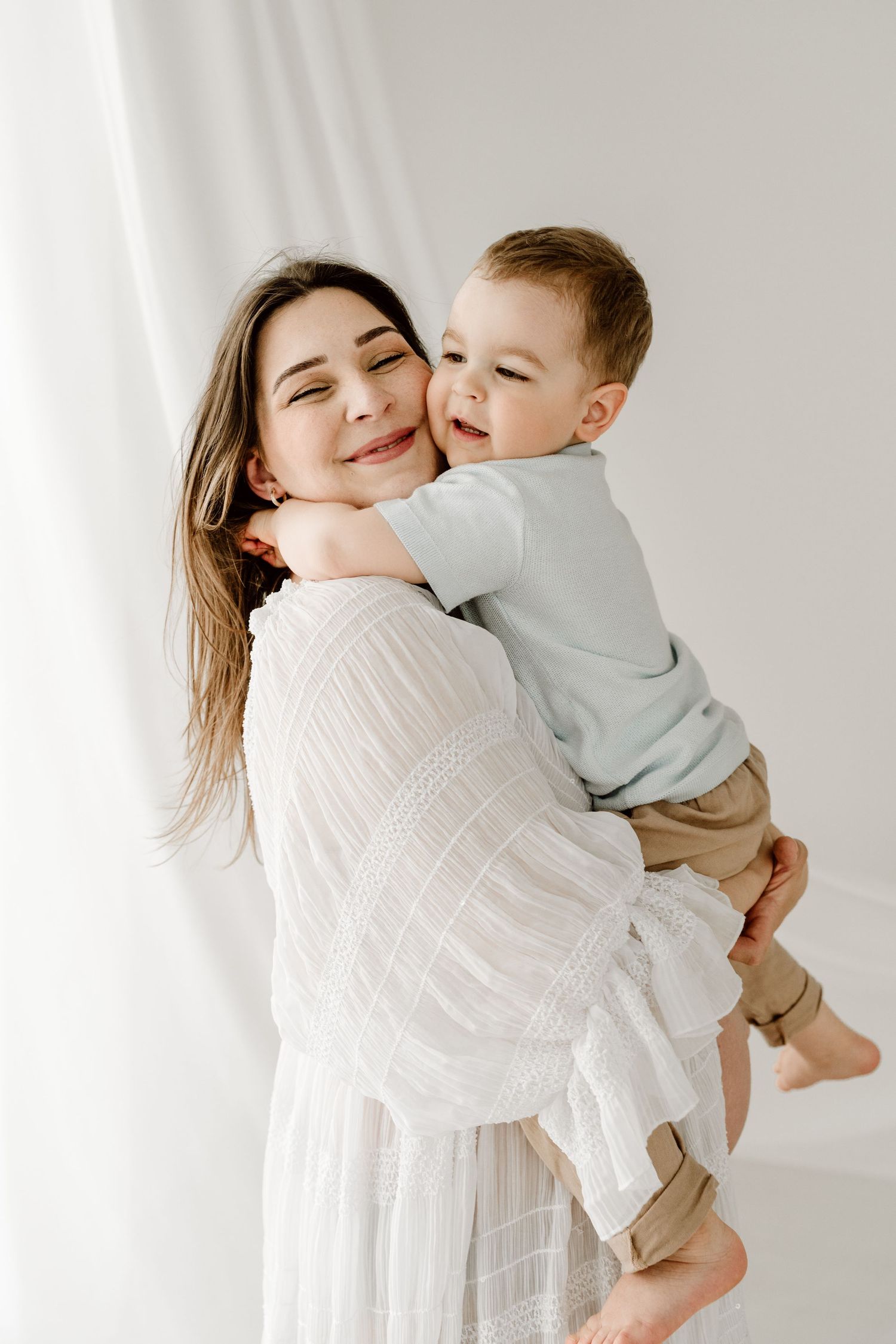A brunette pregnant woman in a white dress smiles while holding her happy toddler son close.