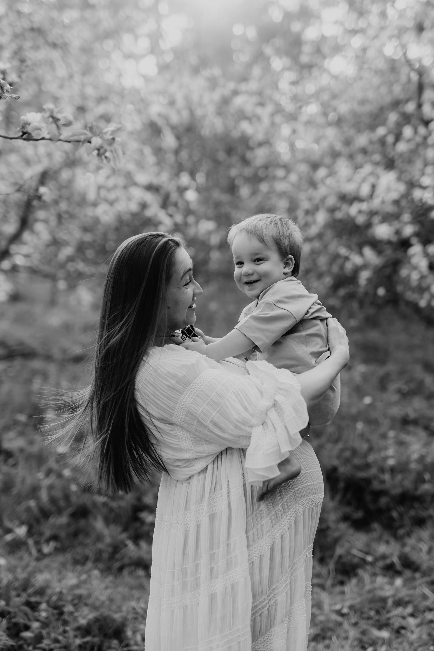 Black and white photo of dark-haired mother in white dress holding smiling baby in blooming orchard.