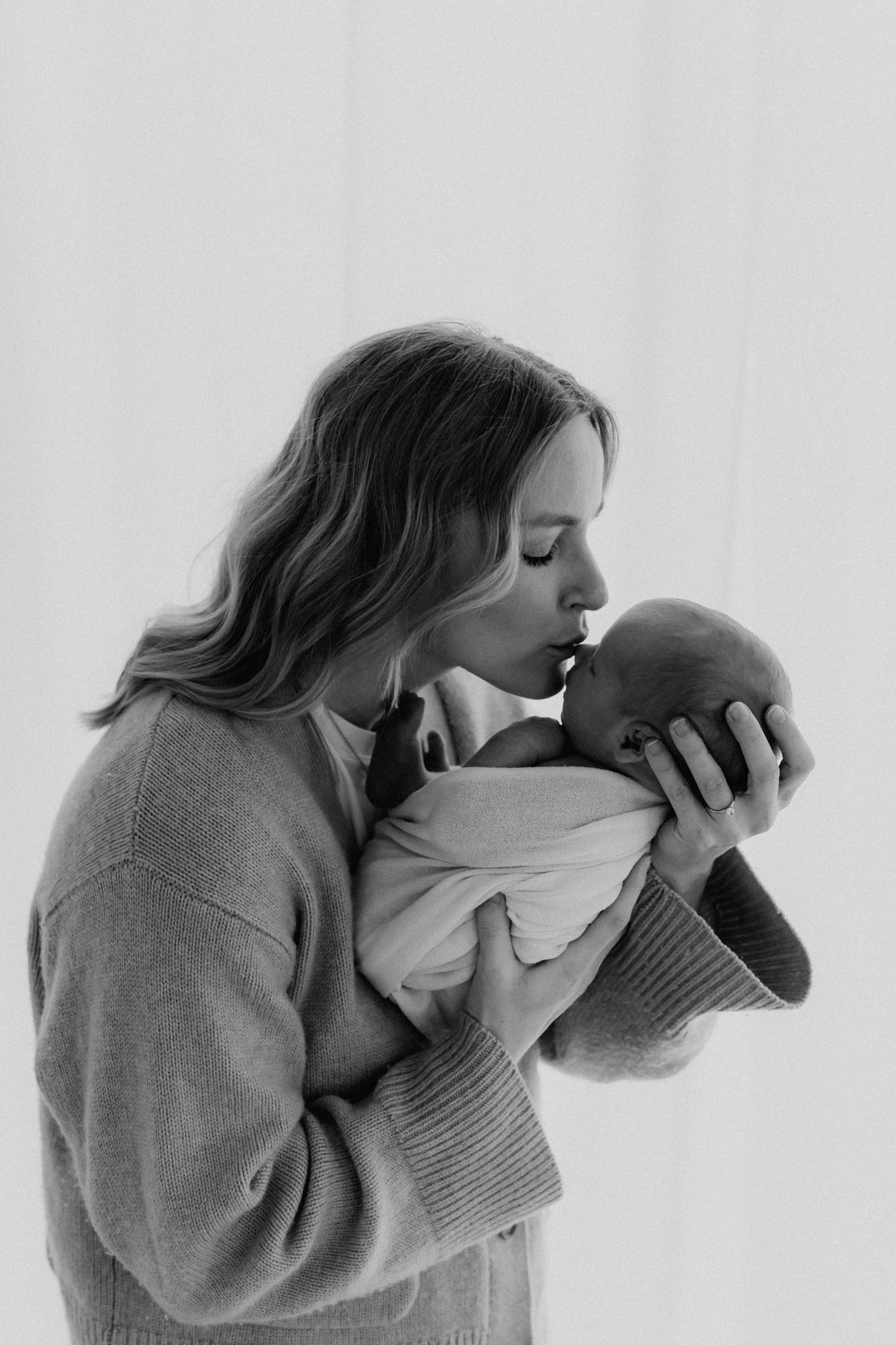 Black and white photo of a mother kissing her sleeping newborn baby wrapped in a white swaddle blanket.