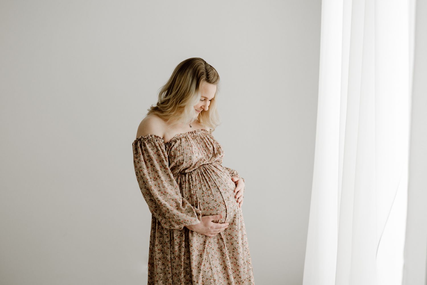Pregnant woman in off-shoulder floral boho dress gently holding baby bump near sheer white curtain in studio.