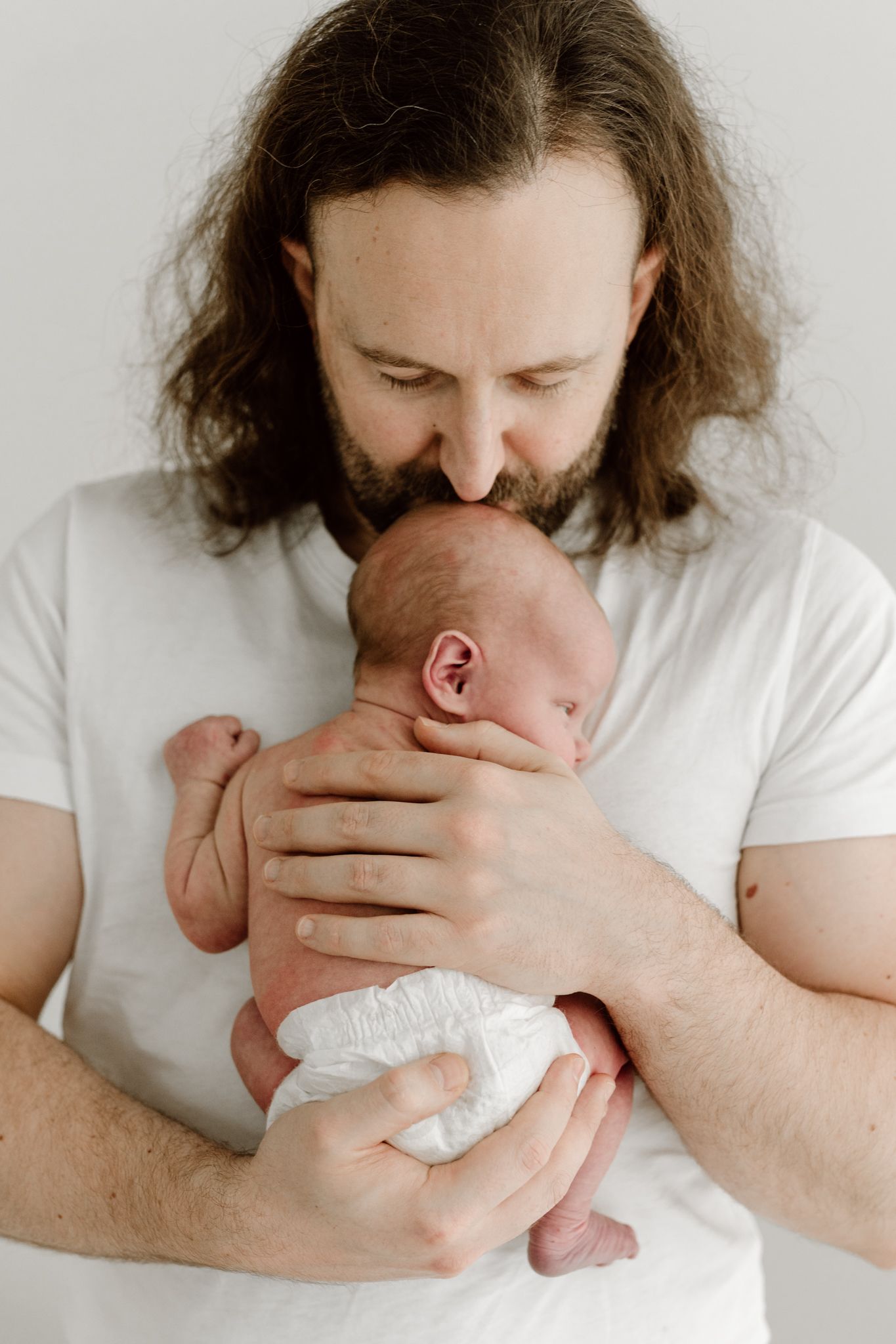 Bearded father with long hair gently kissing his newborn baby's head in a white studio.