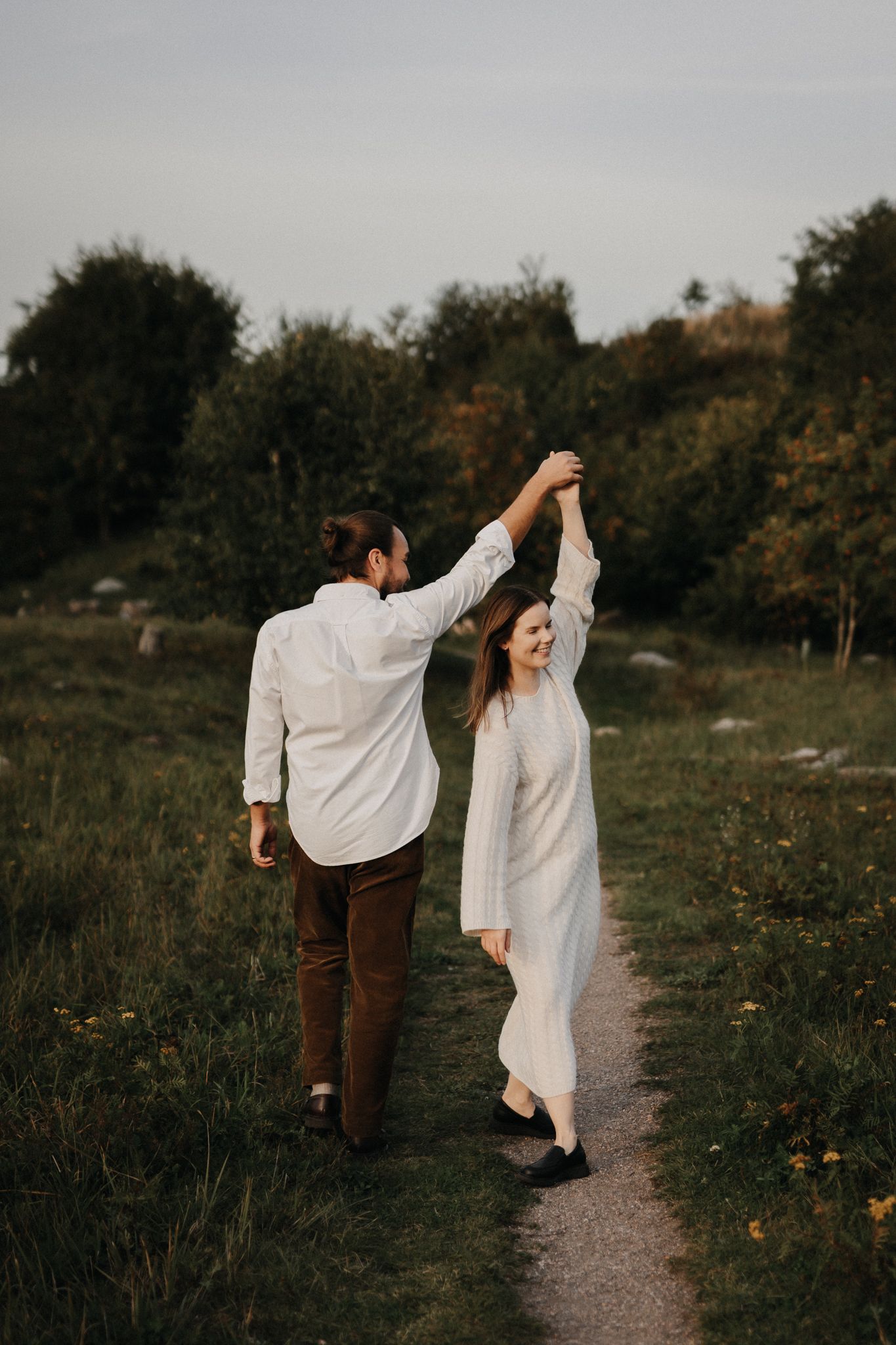 A joyful couple in white holding hands and dancing on a nature path at dusk.
