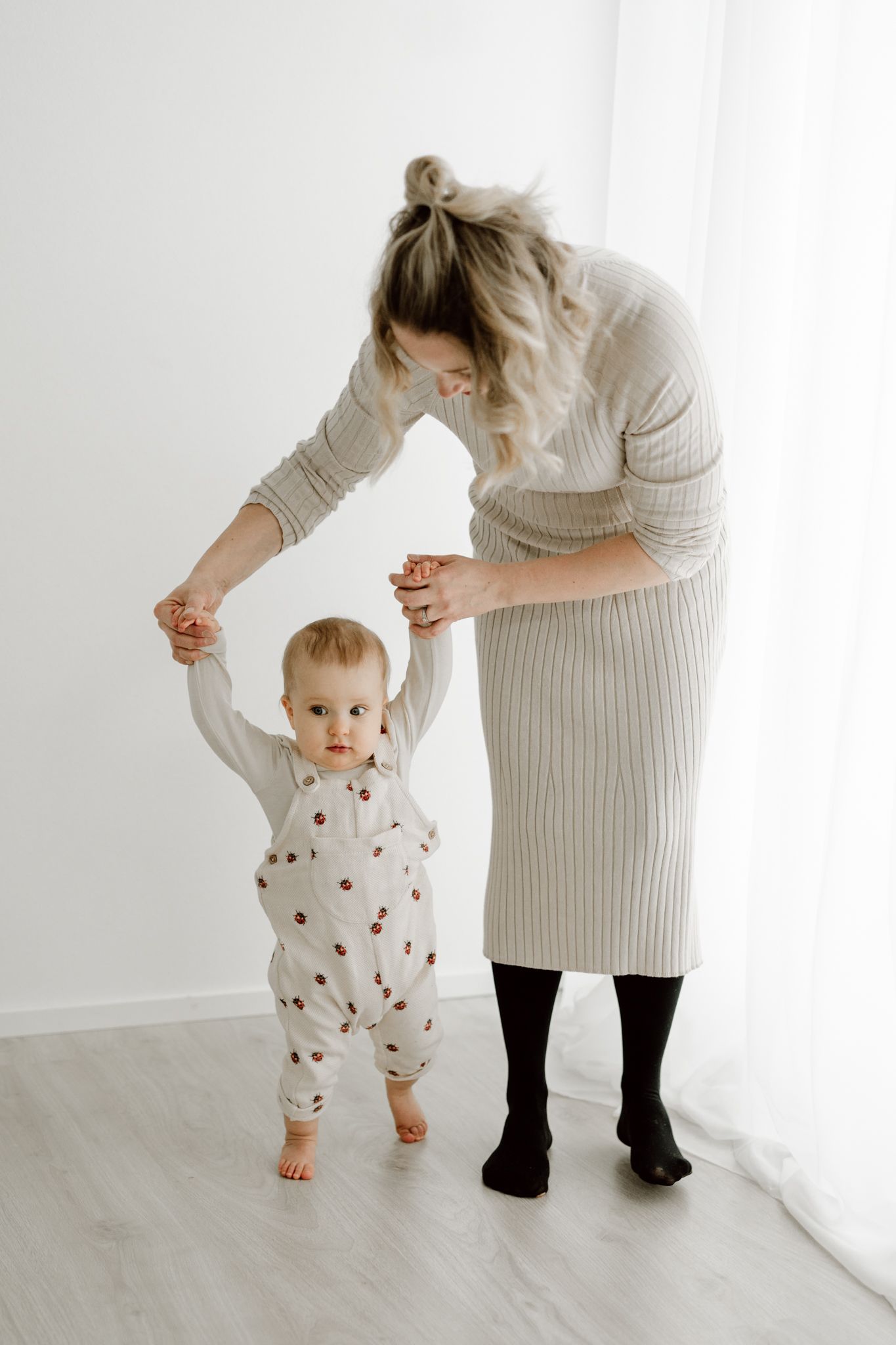 Mother in a ribbed dress helping her baby take first steps near a bright window.