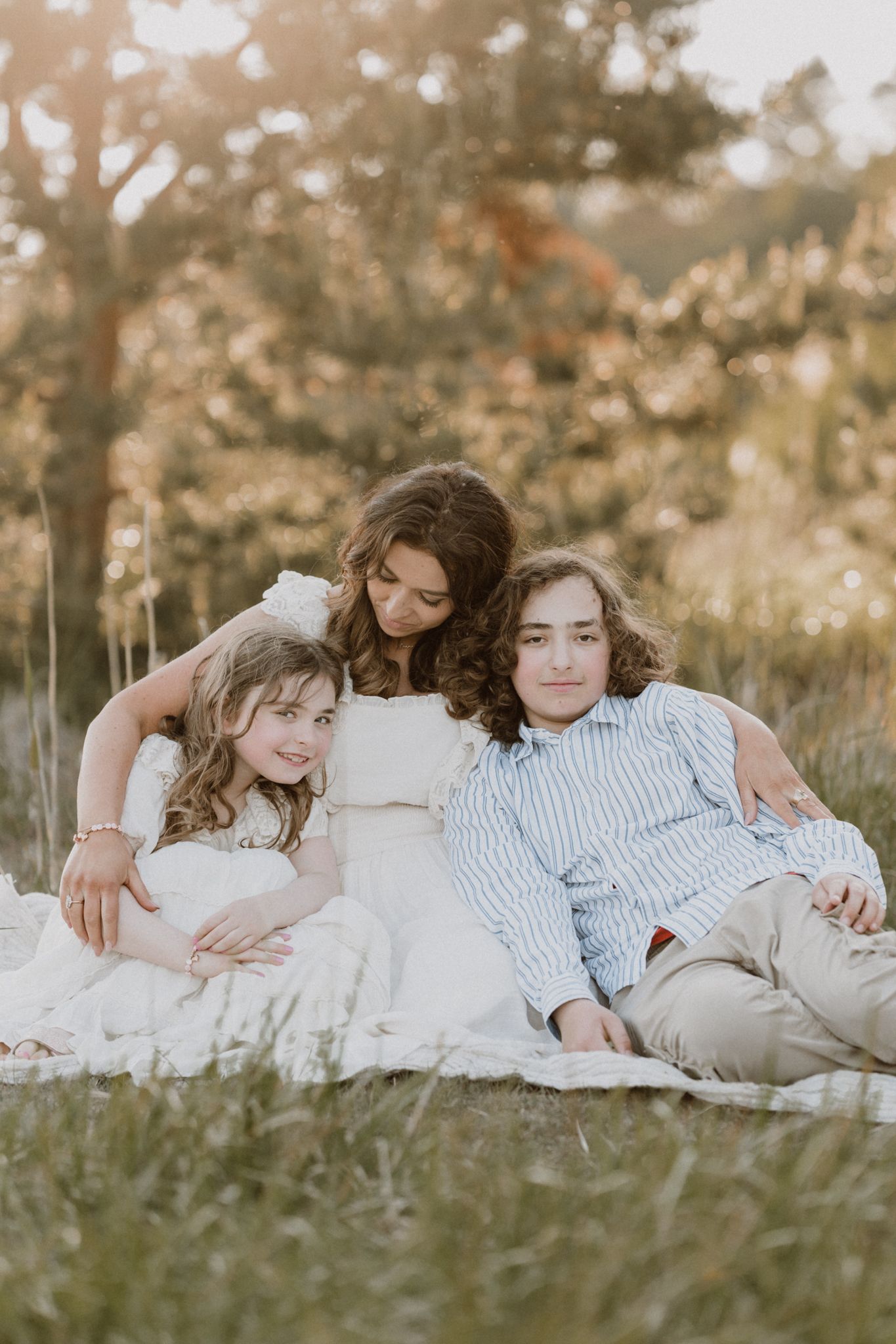 A mother and two children relax together on a blanket in a sun-drenched grassy field.