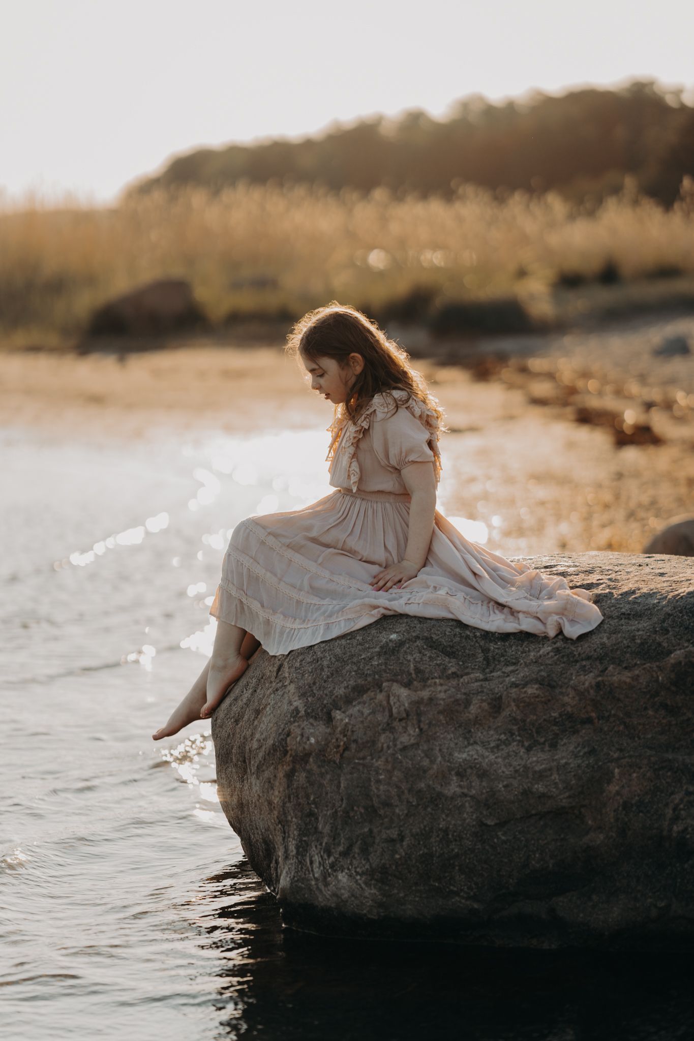 A young girl in a light dress sits barefoot on a rock by the water at sunset.