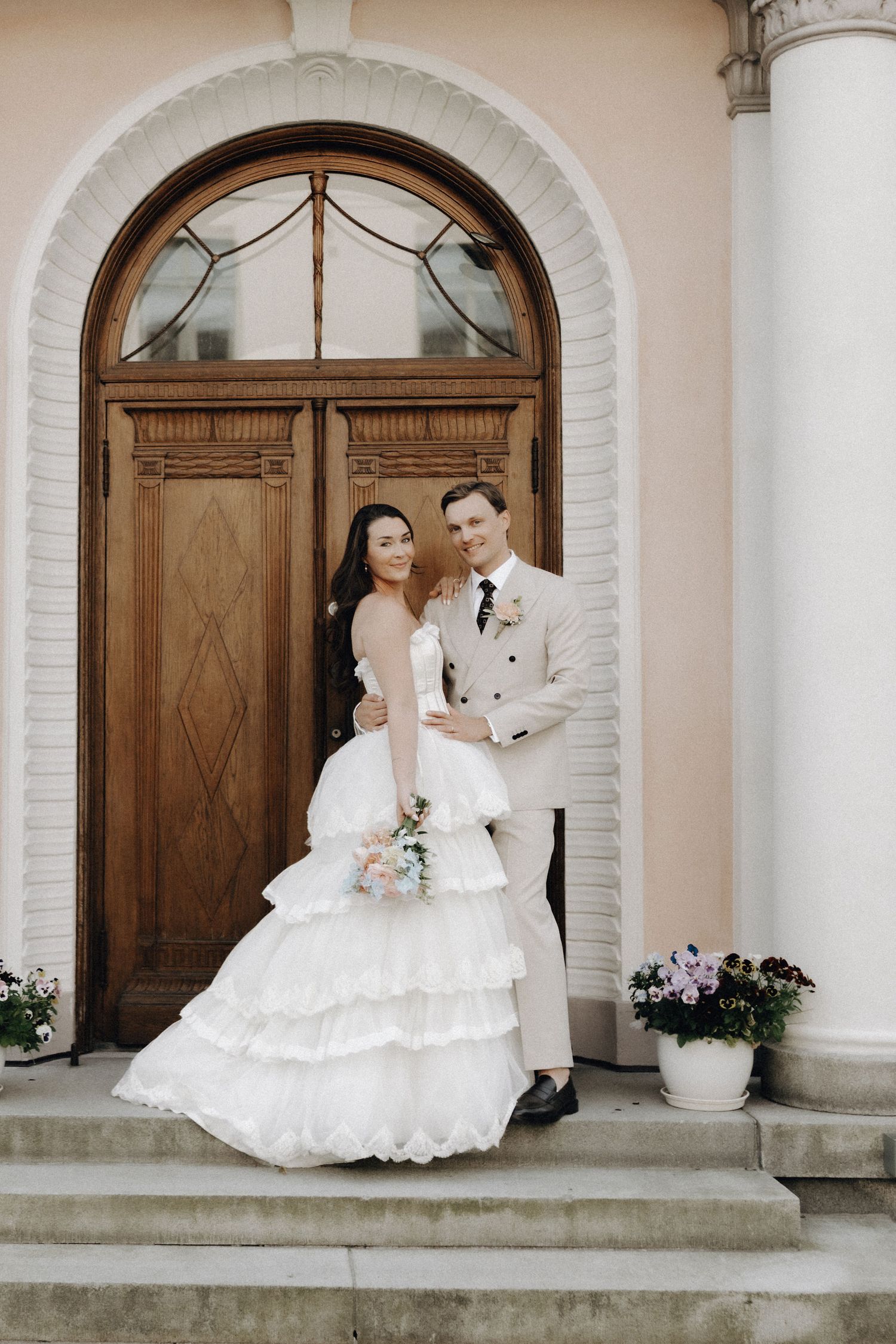 Bride and groom pose together on steps before a grand arched wooden door of an elegant building.