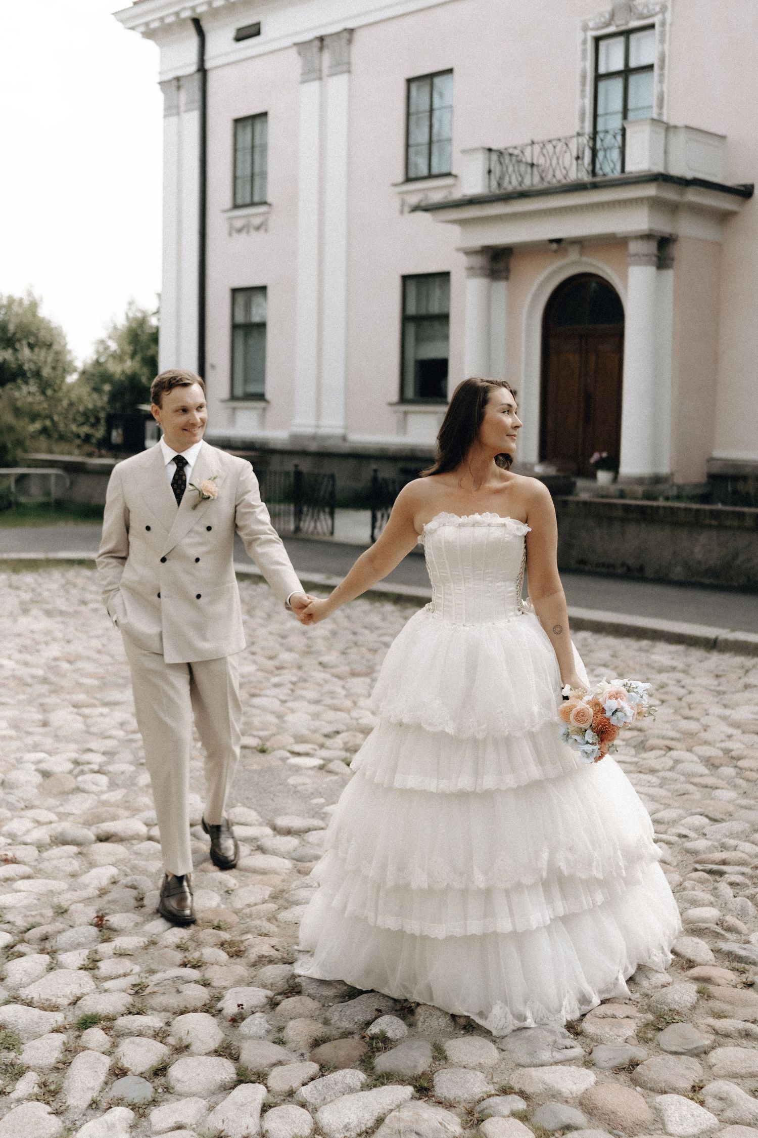 Bride and groom holding hands walking on cobblestone path before a historic building.