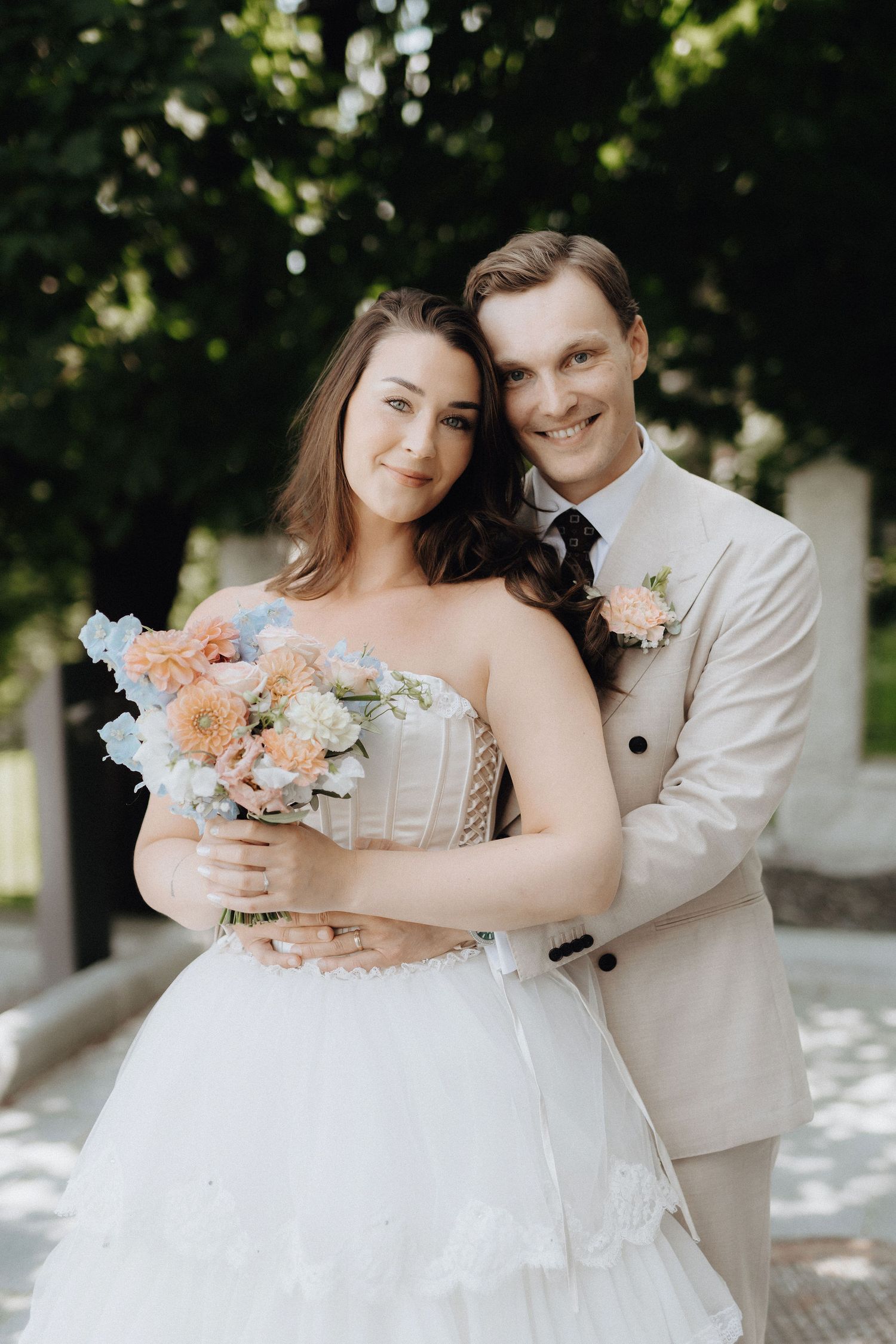Happy couple posing together outdoors, bride in strapless white gown holds pastel bouquet as groom smiles warmly.