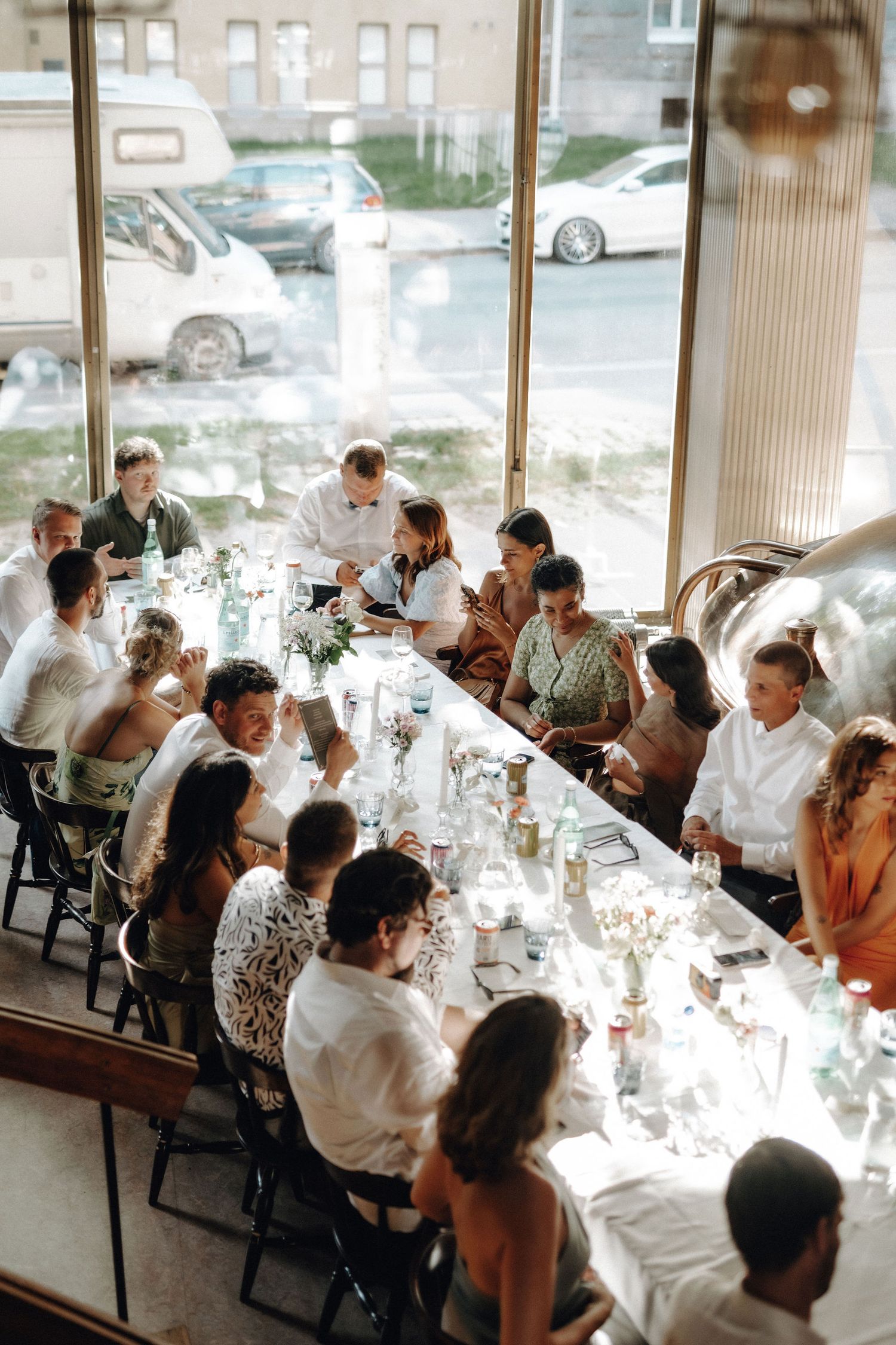 Aerial view of wedding guests gathered around long decorated reception table near large windows.
