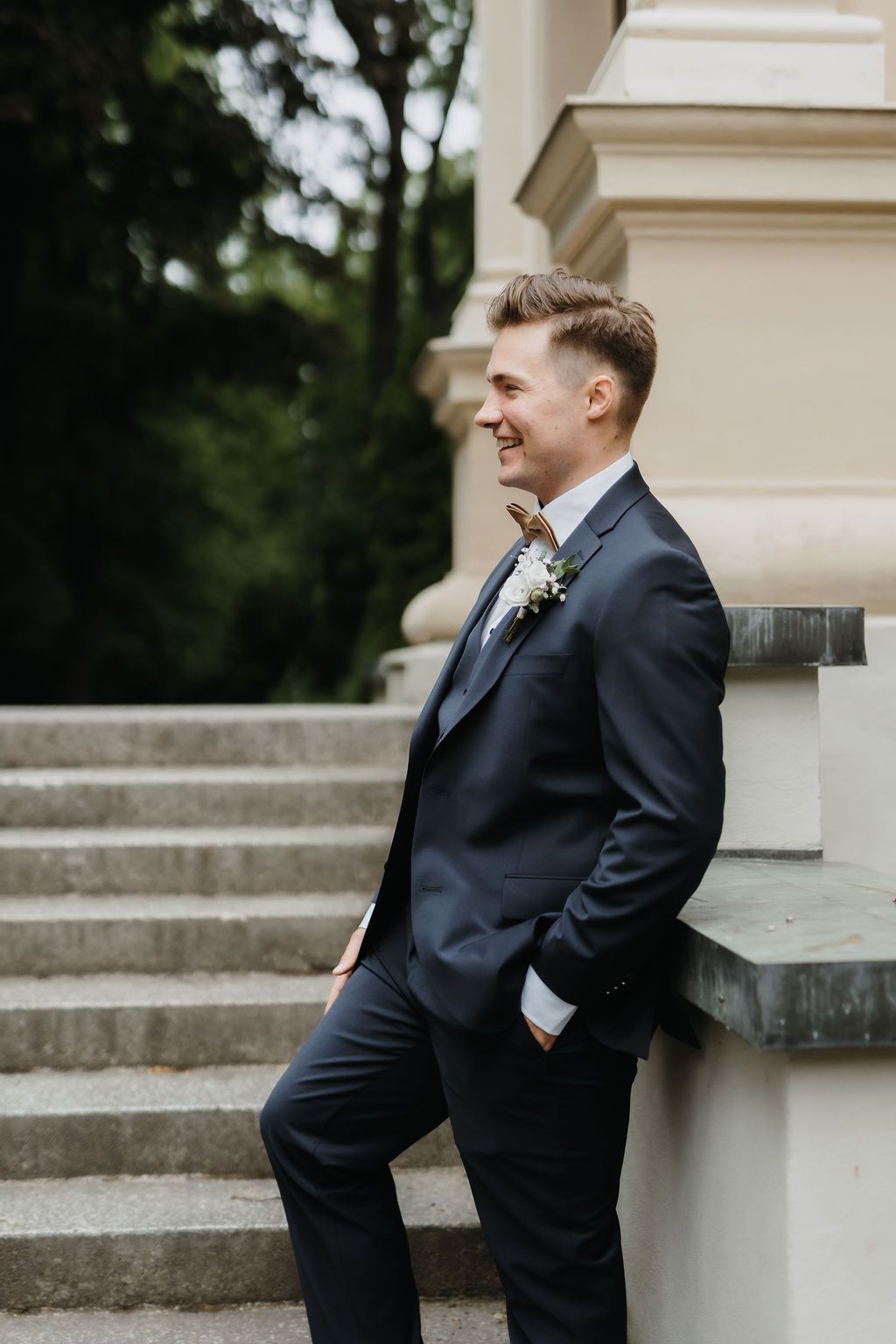 Smiling groom in navy suit with boutonniere leaning against stone pillar at outdoor wedding venue.