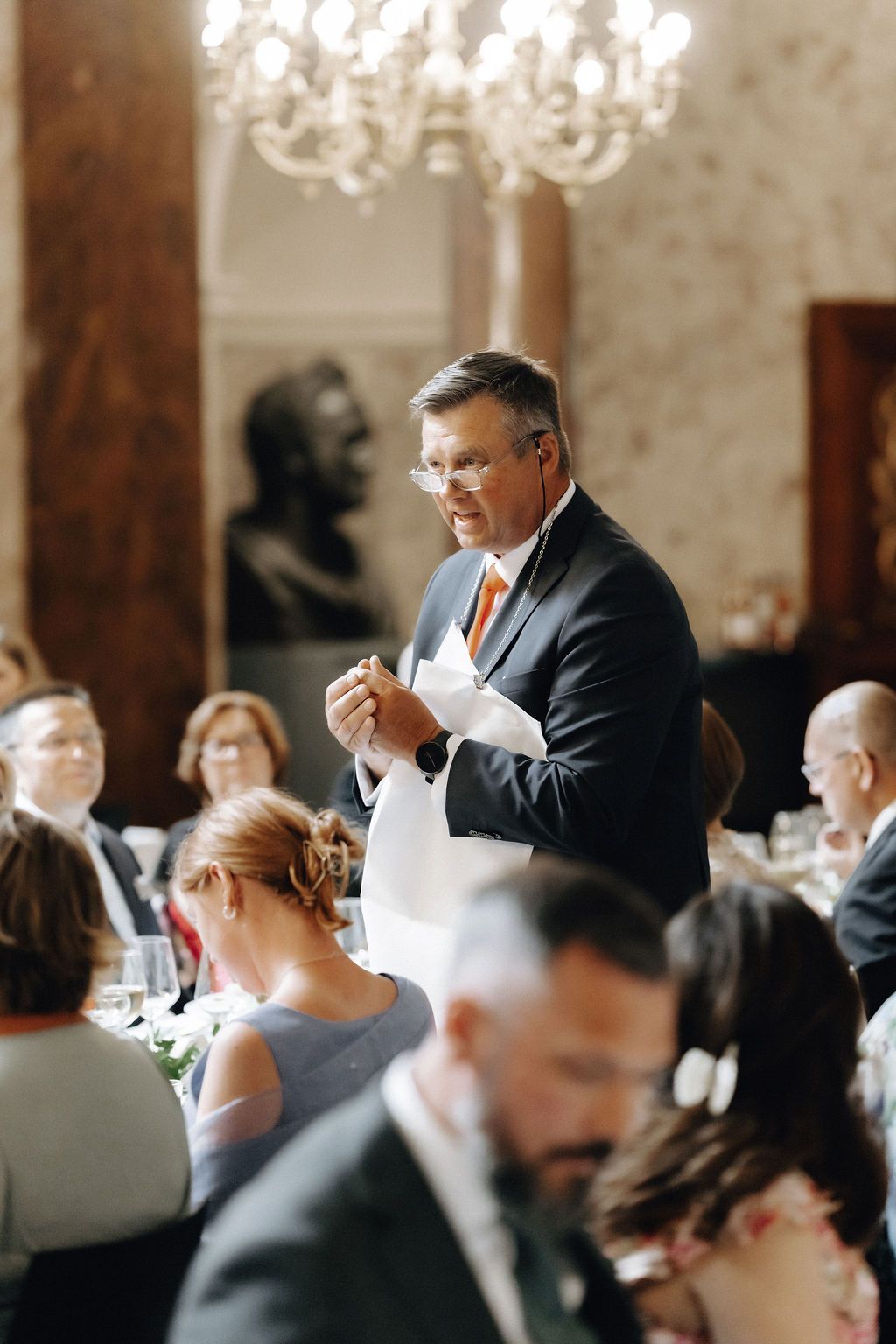 Man in suit with orange tie delivers speech at elegant wedding reception dinner.