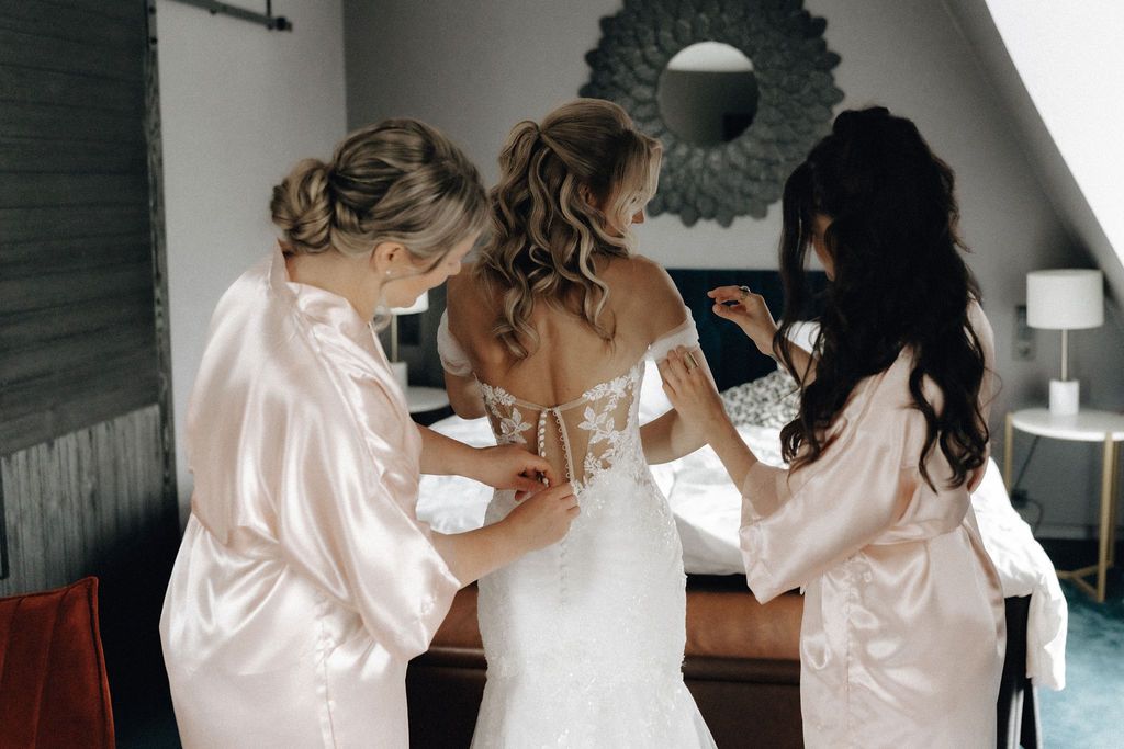 Two bridesmaids in satin robes helping bride button her lace wedding gown before ceremony.