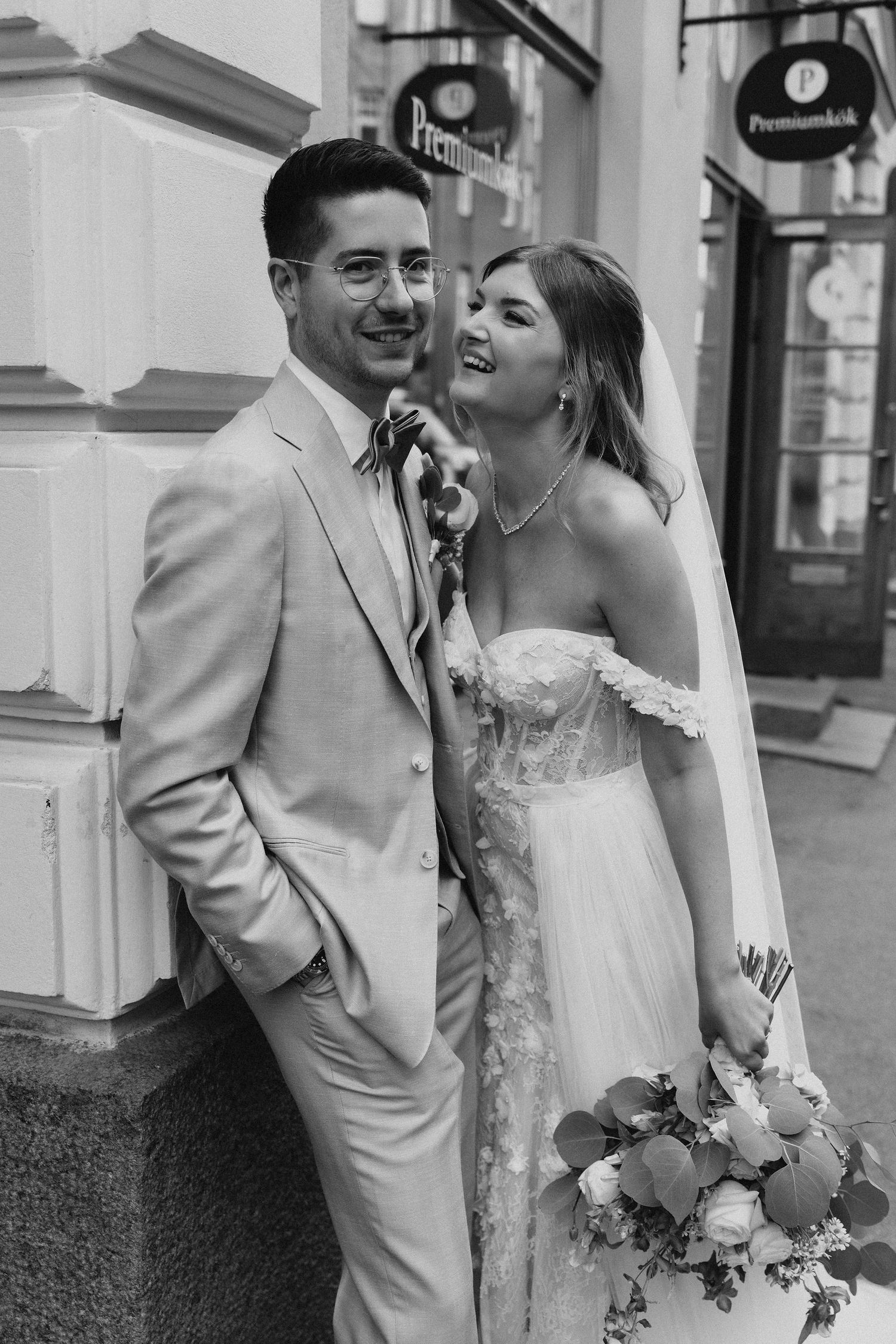 Laughing bride leans on groom in beige suit outdoors in black and white wedding portrait.