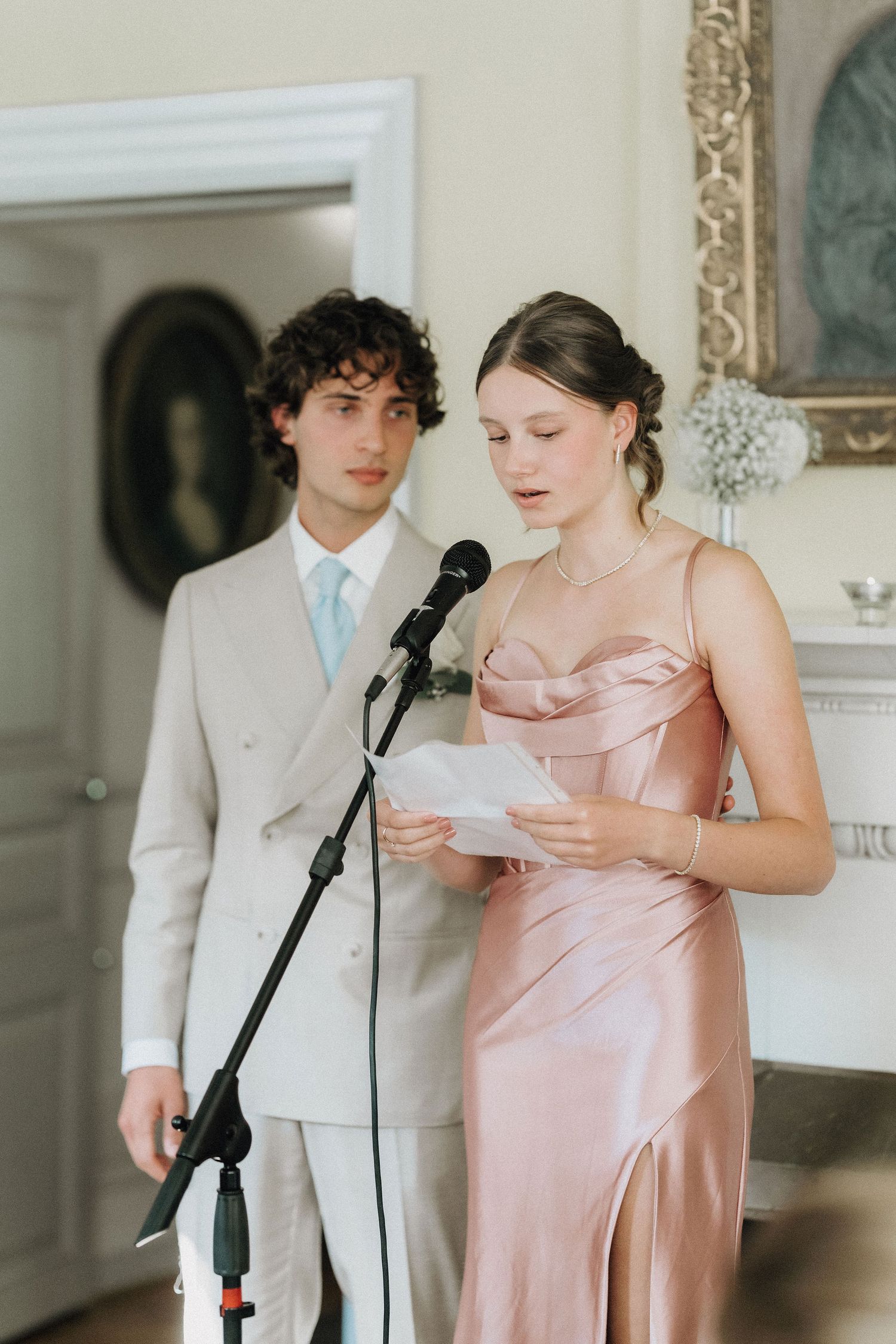 Young woman in pink satin dress giving a wedding speech at microphone beside man in beige suit.