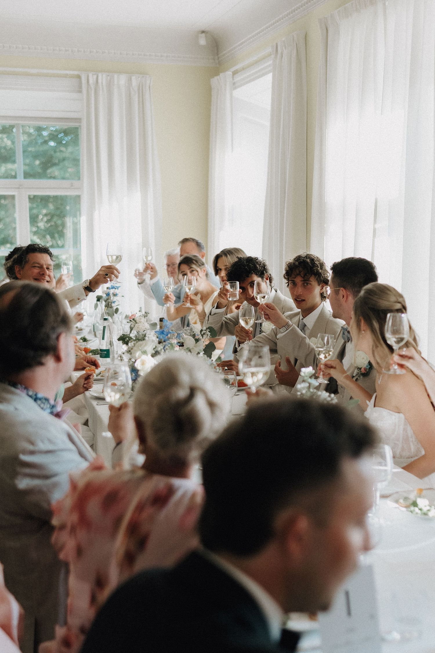 Wedding guests raising glasses in a celebratory toast at a bright, elegantly decorated reception dinner table.