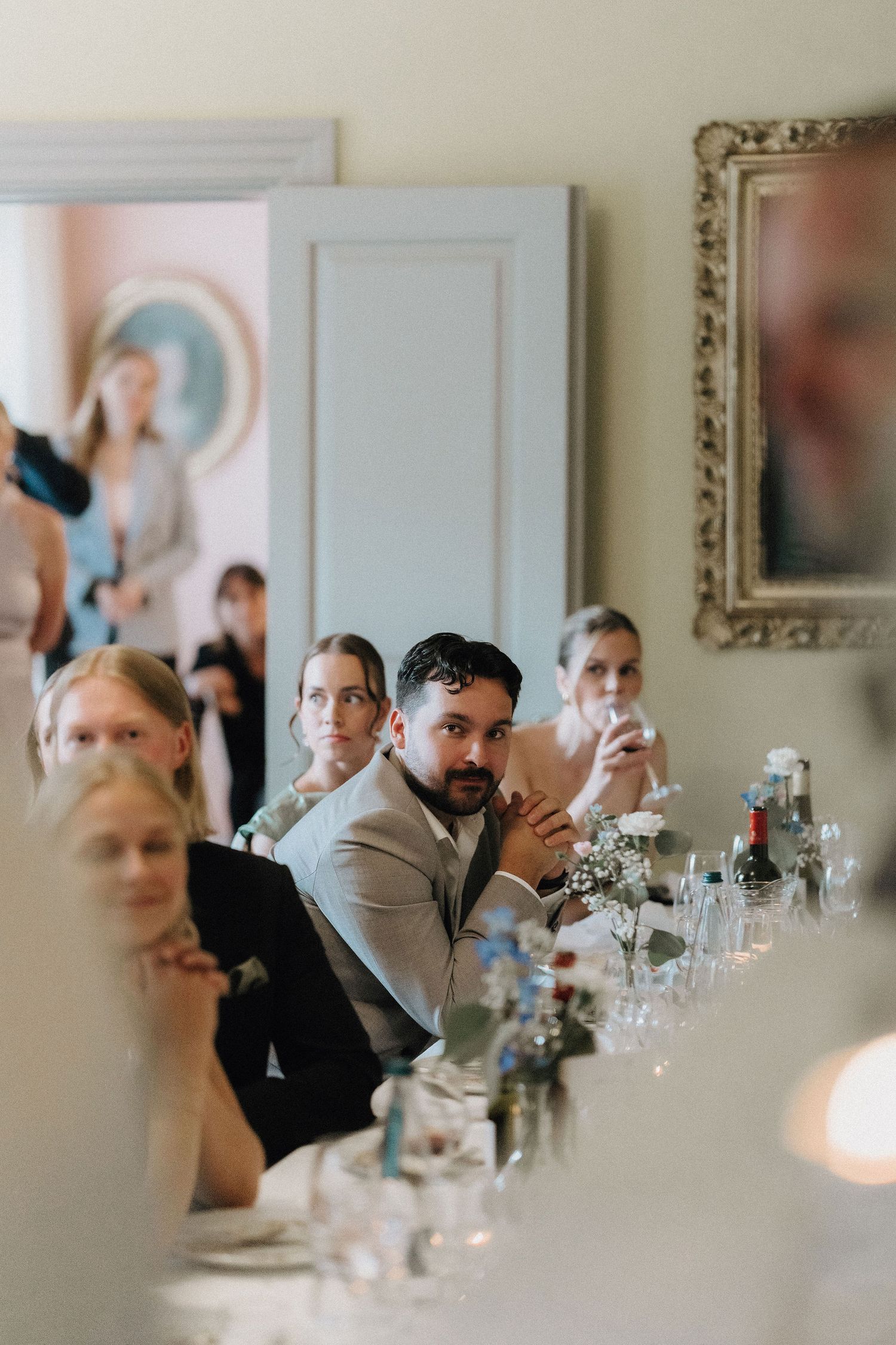 Wedding guests seated at long reception table listening attentively to speeches in elegant venue.