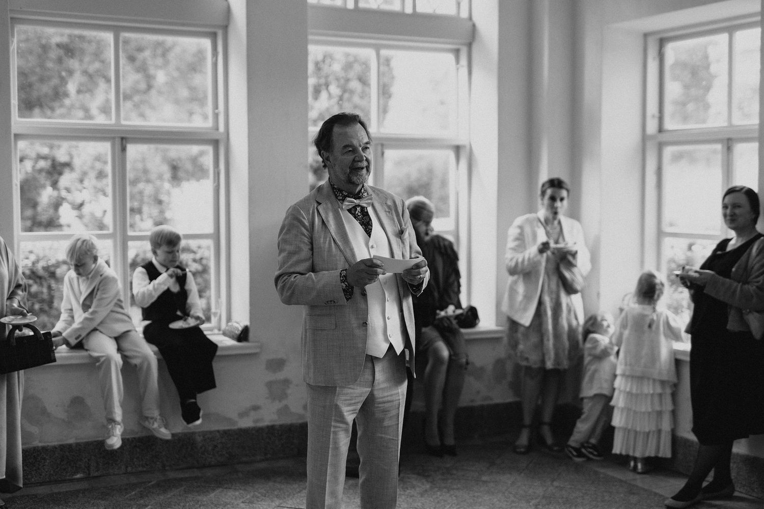 Black and white photo of older man in suit speaking to wedding guests in elegant venue.