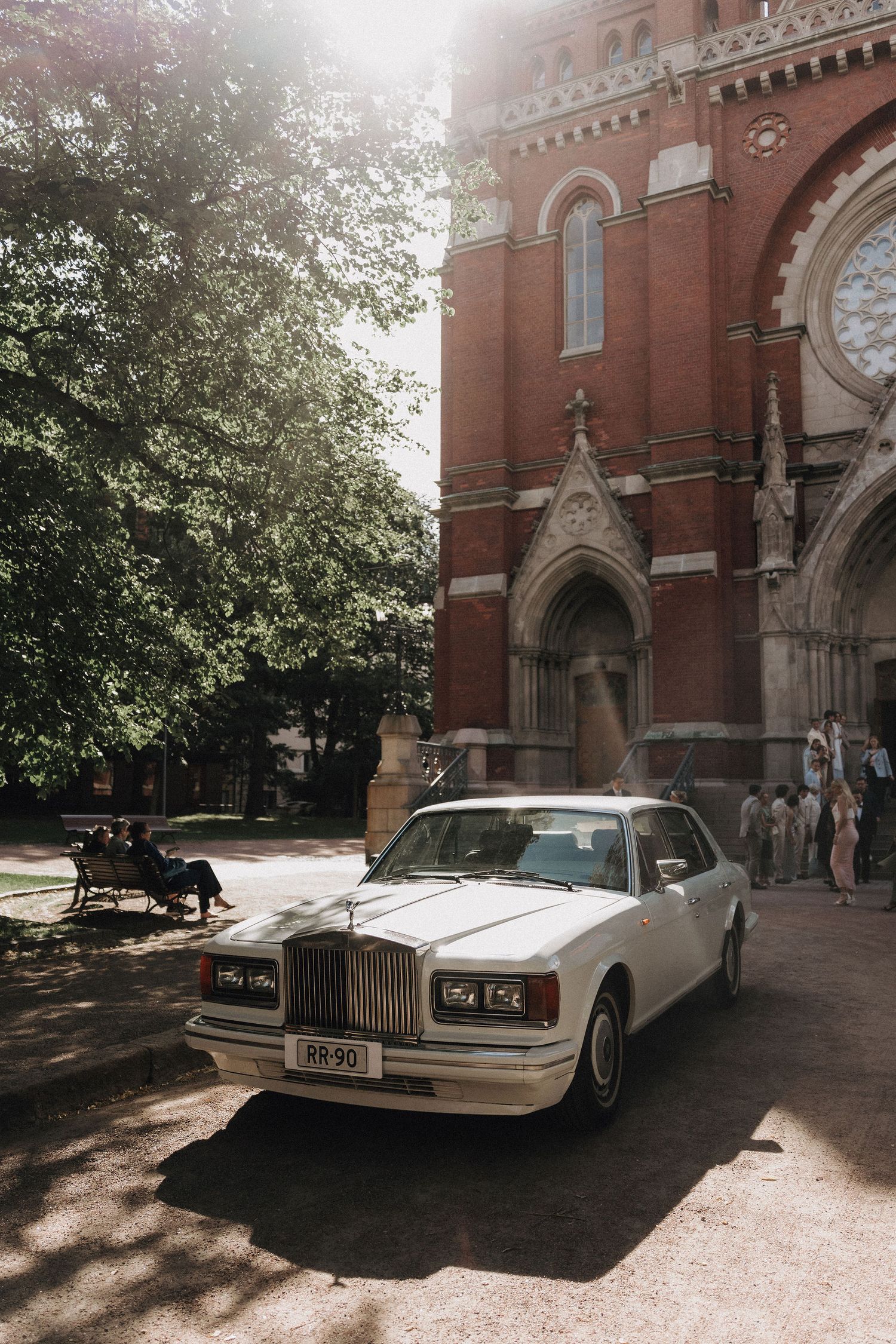 White Rolls-Royce wedding car parked outside a Gothic red brick church on a sunny day.