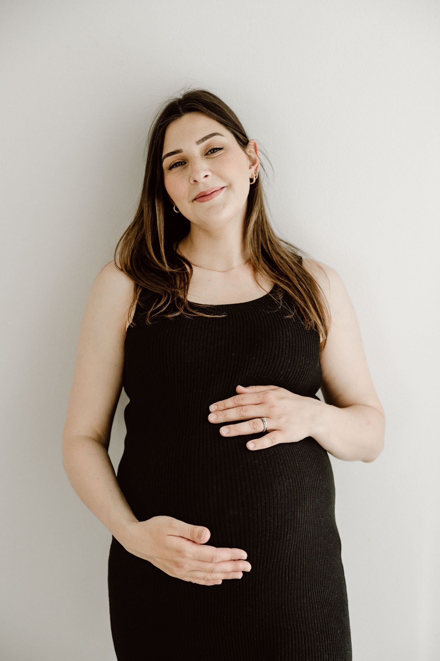 Pregnant woman in black dress cradling baby bump with both hands against white wall.