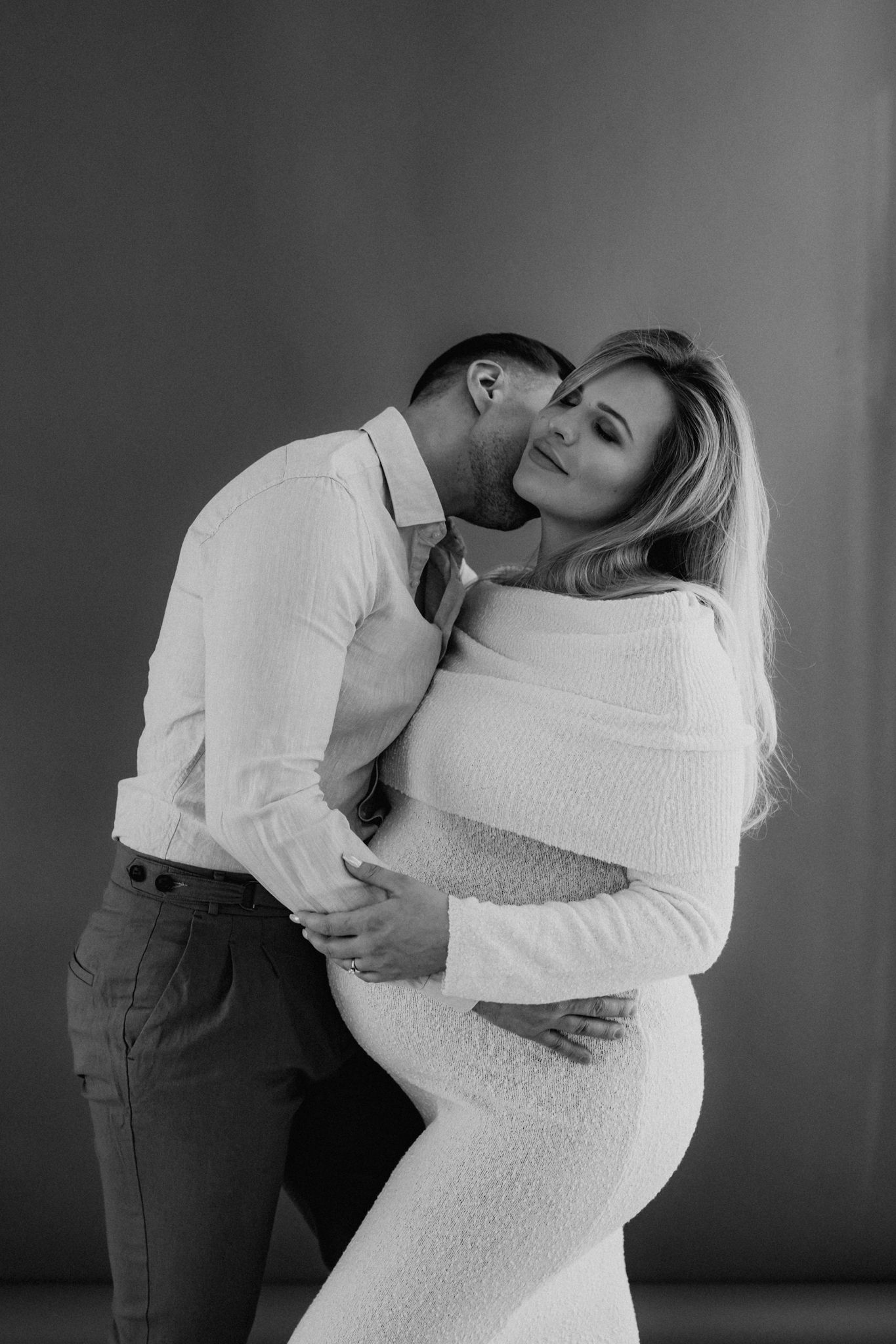 Black and white maternity portrait of couple embracing, man kissing pregnant woman in white knit dress.