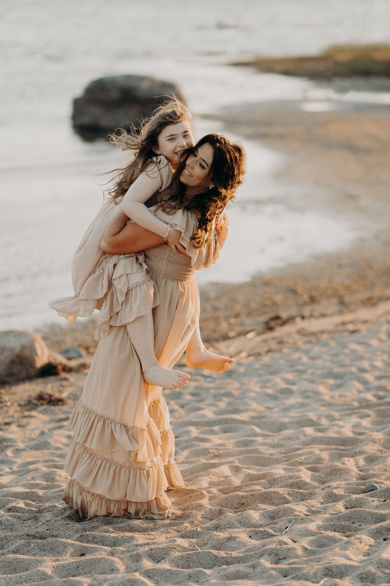 Woman in beige dress giving girl piggyback ride on sandy beach at golden hour sunset.