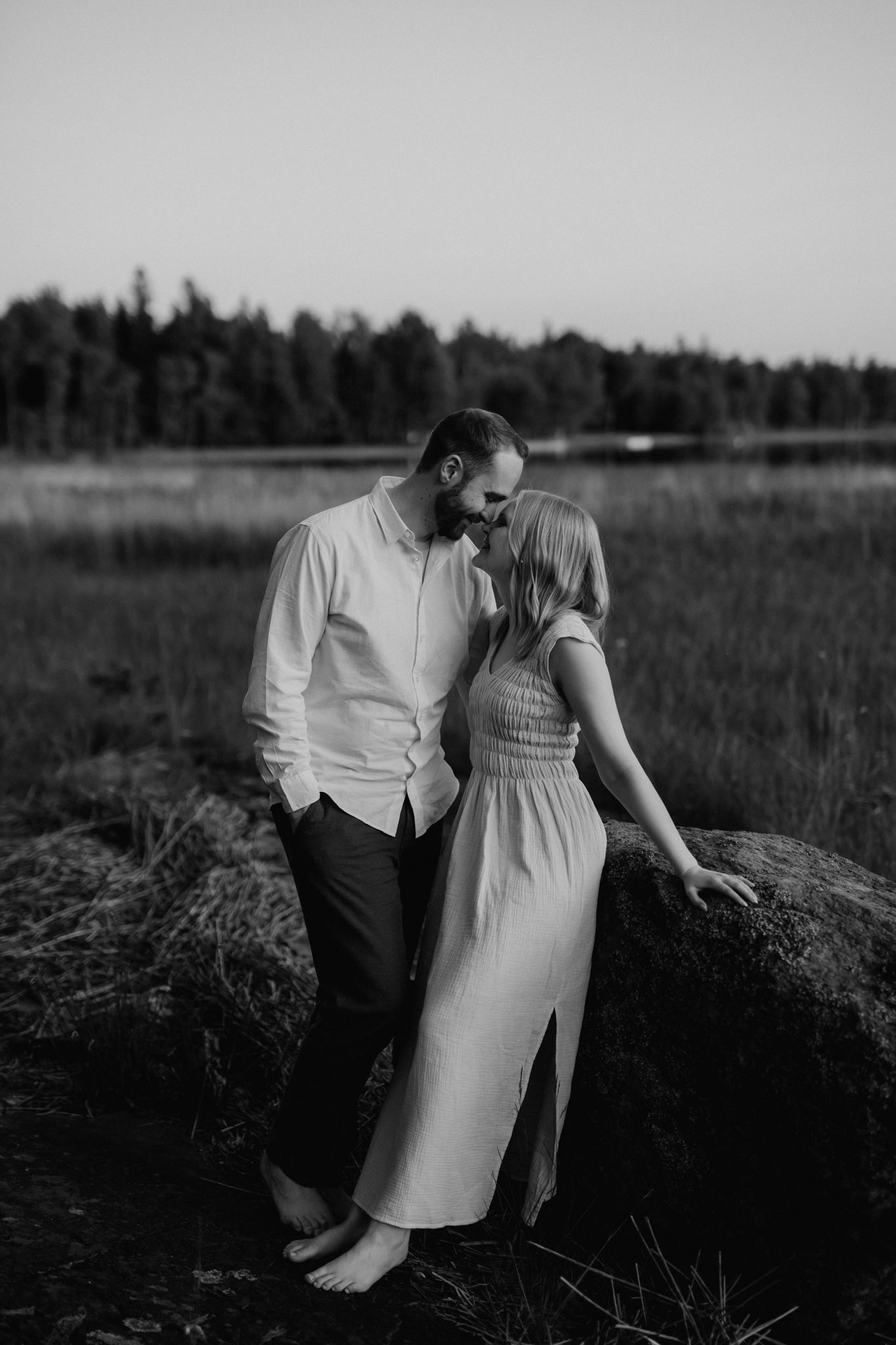 Black and white photo of a couple leaning against a boulder, sharing an intimate moment at dusk.