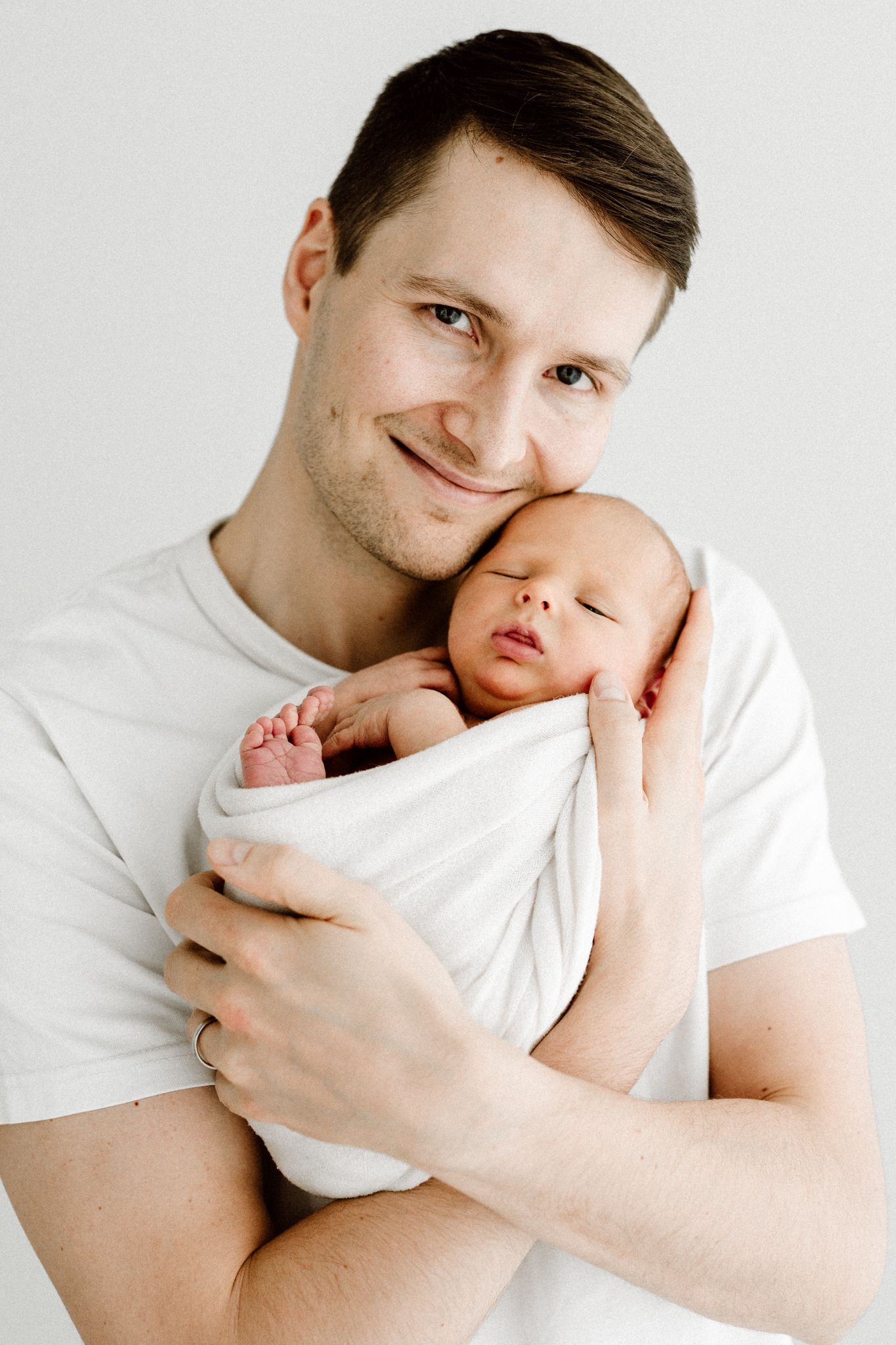 Happy father in white t-shirt cradling his sleeping swaddled newborn baby against his cheek.