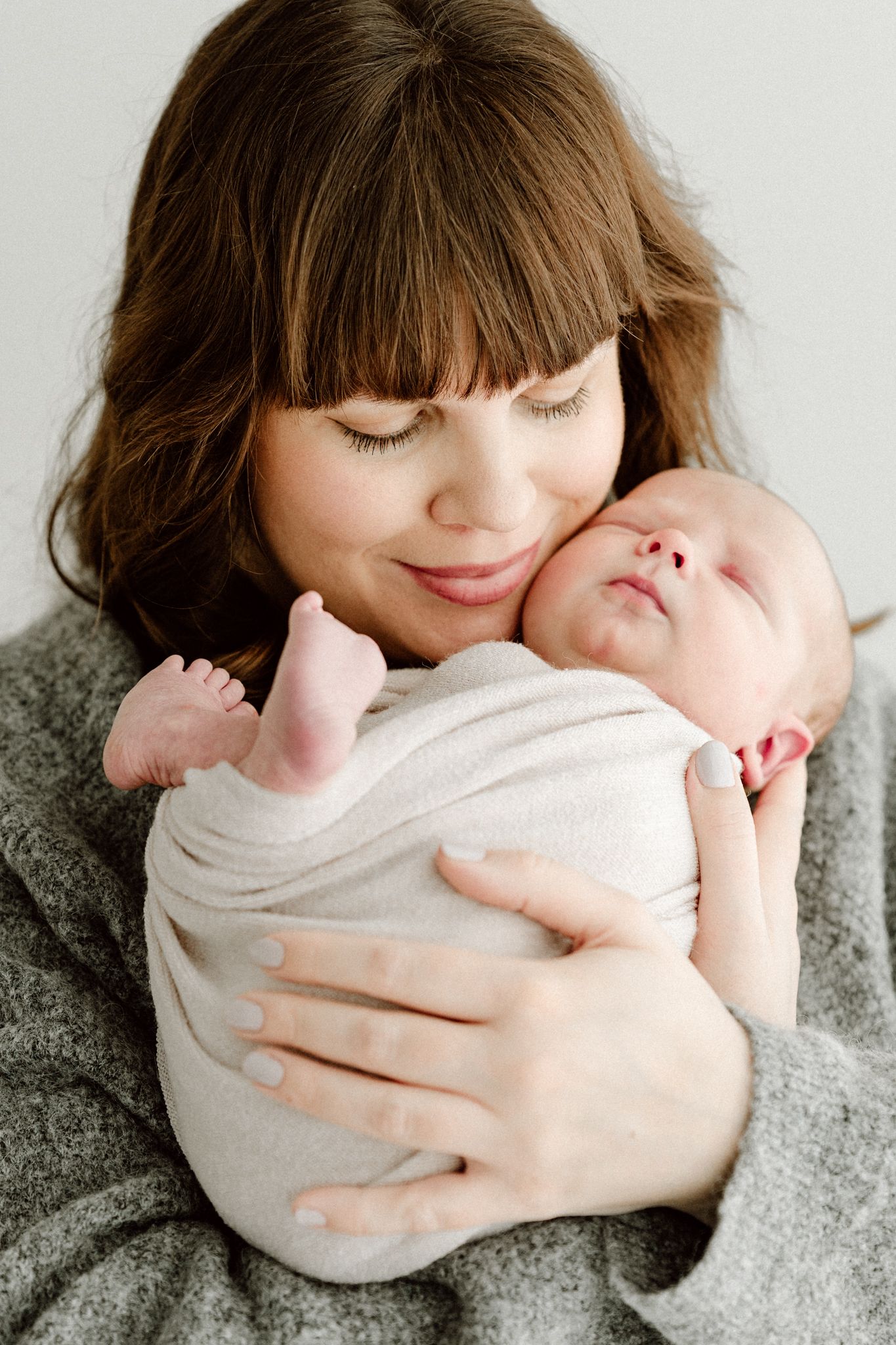 Brunette mother with bangs smiling while gently holding swaddled newborn baby close to her face.