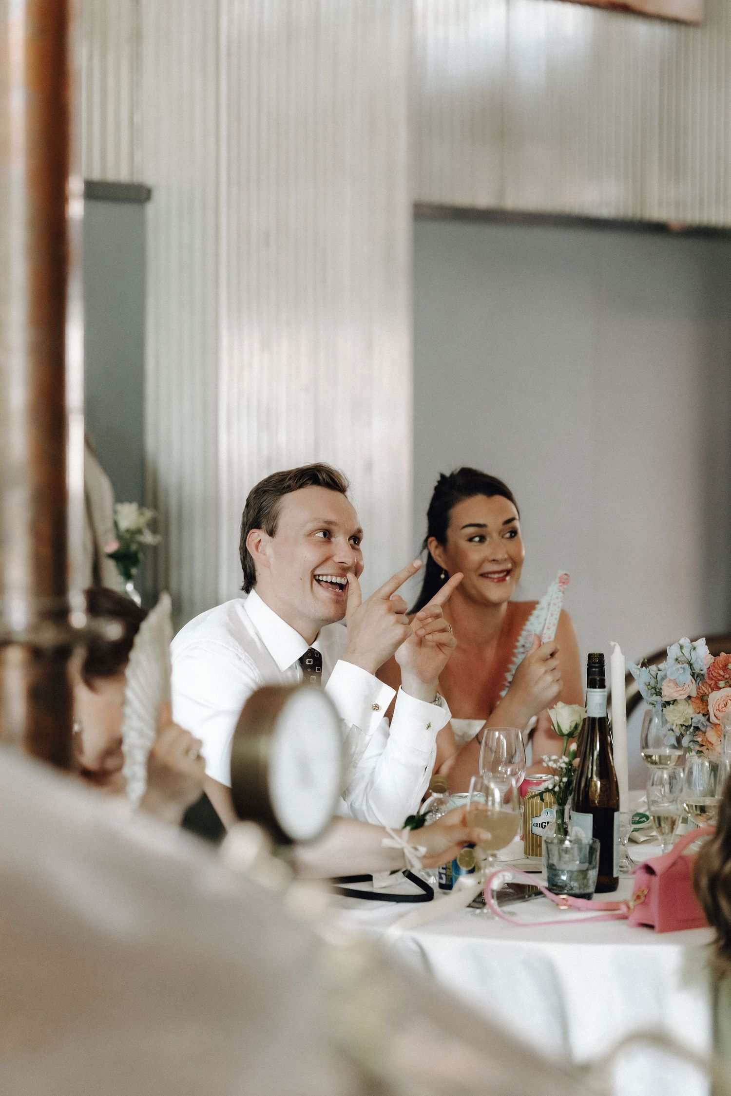 Laughing bride and groom seated at wedding reception table with wine bottles and floral centerpieces.