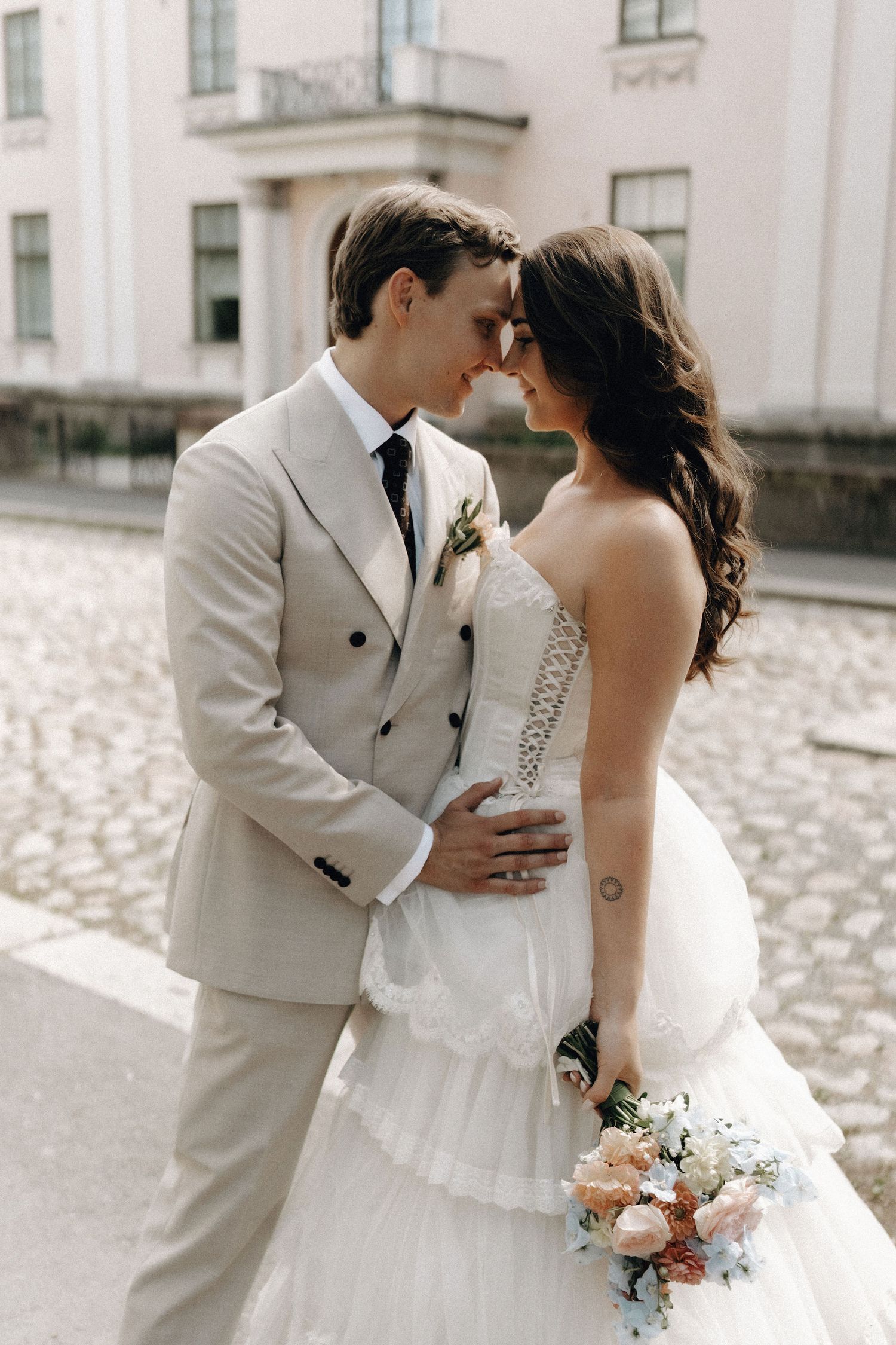 Bride and groom touching foreheads tenderly on cobblestone street outside a historic building.