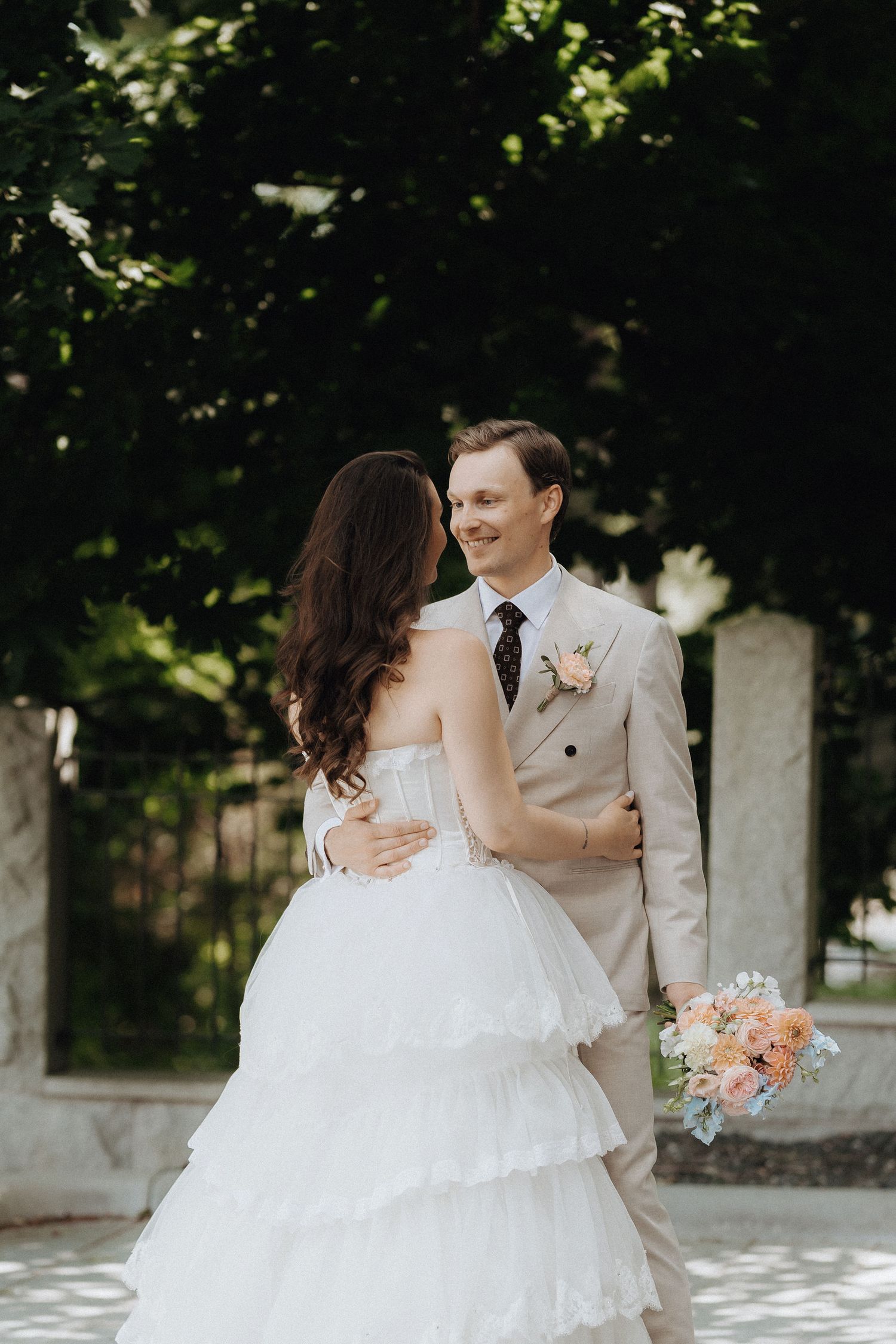 Wedding couple embracing outdoors, bride in layered white dress, groom in beige suit holding floral bouquet.
