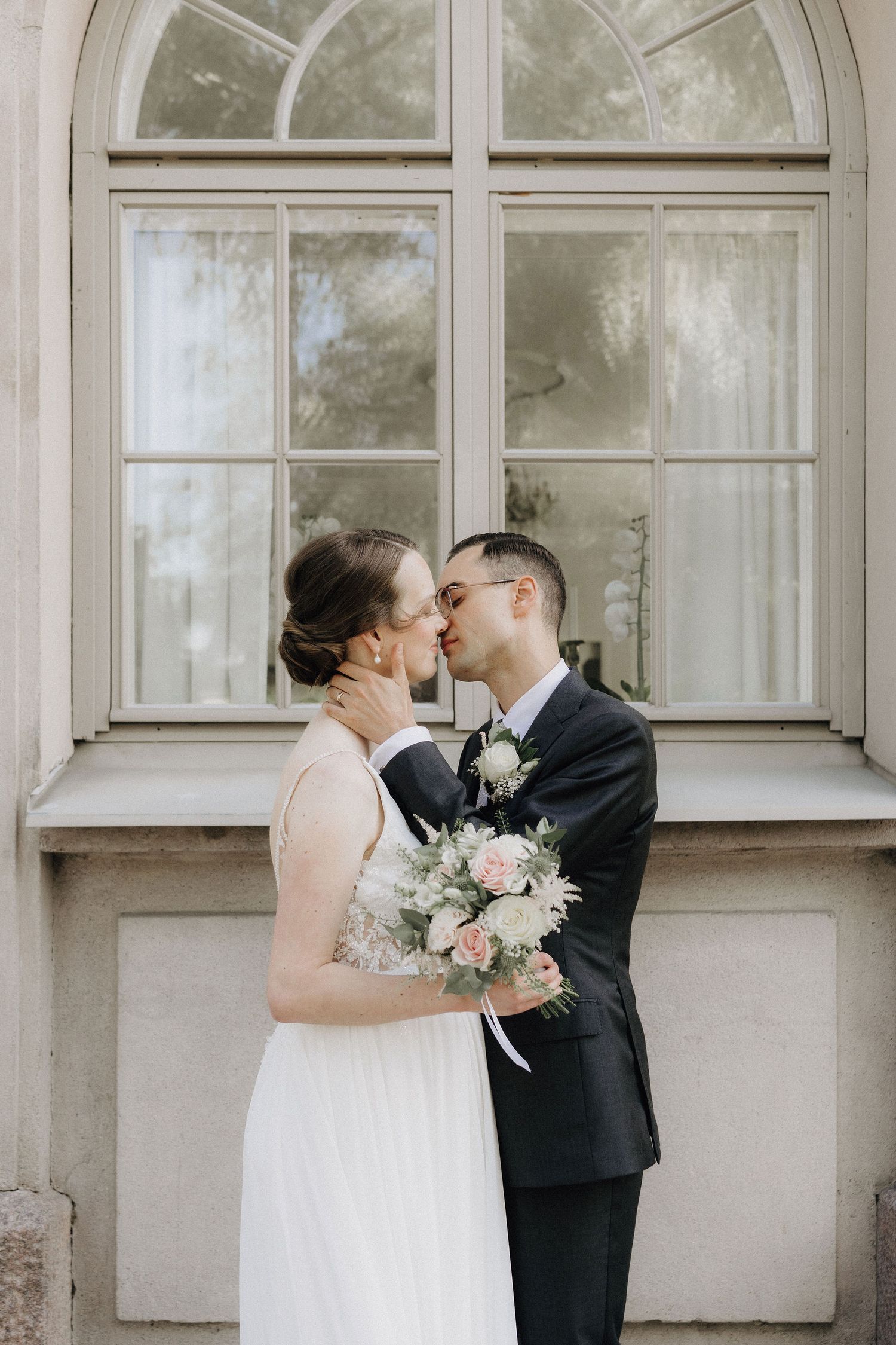Bride and groom share a kiss in front of an arched window, bride holding a soft pastel bouquet.