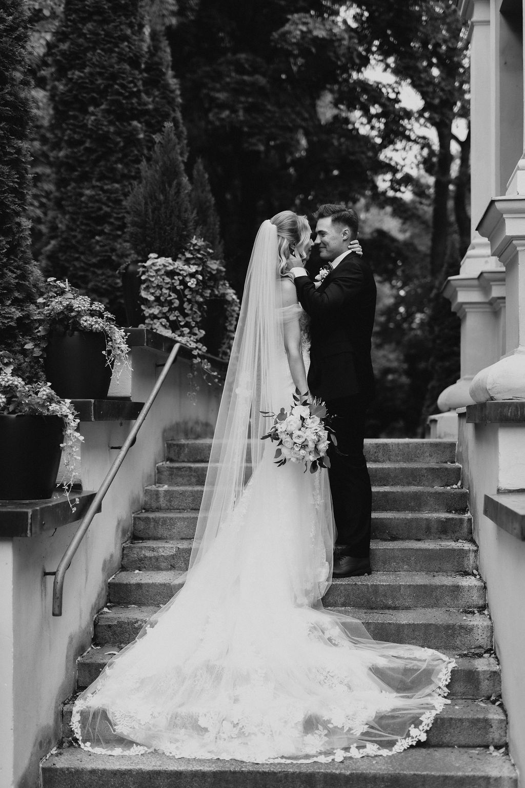 Black and white wedding portrait of couple embracing on stone staircase with long lace cathedral veil trailing.