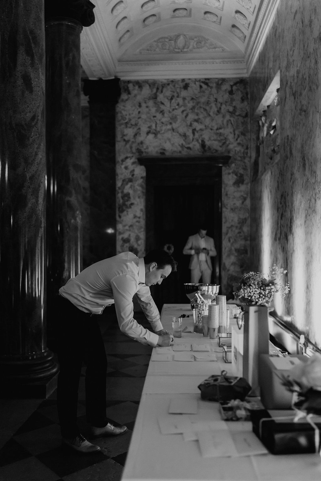 Black and white photo of a man arranging place cards on a table in an ornate venue hallway.