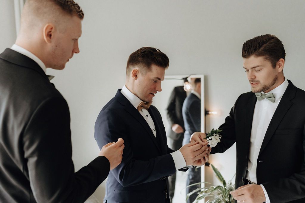 Groomsmen in black suits pinning boutonniere on groom's lapel before wedding.