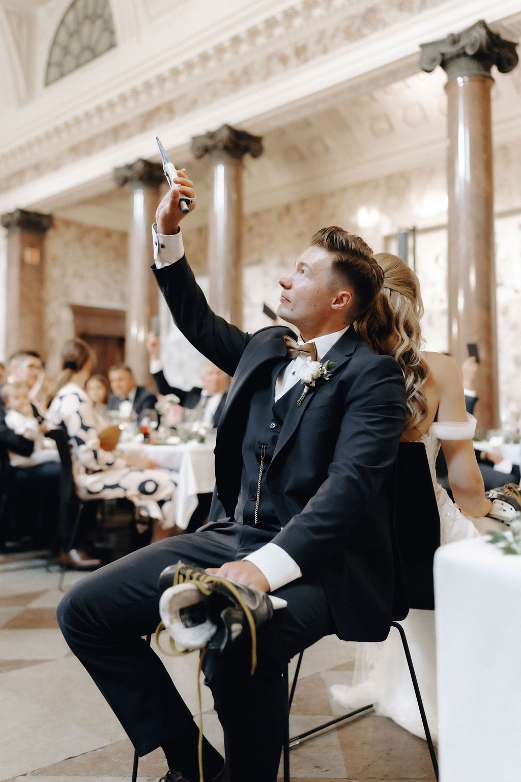 Groom takes a selfie with bride at sweetheart table in ornate wedding reception venue.