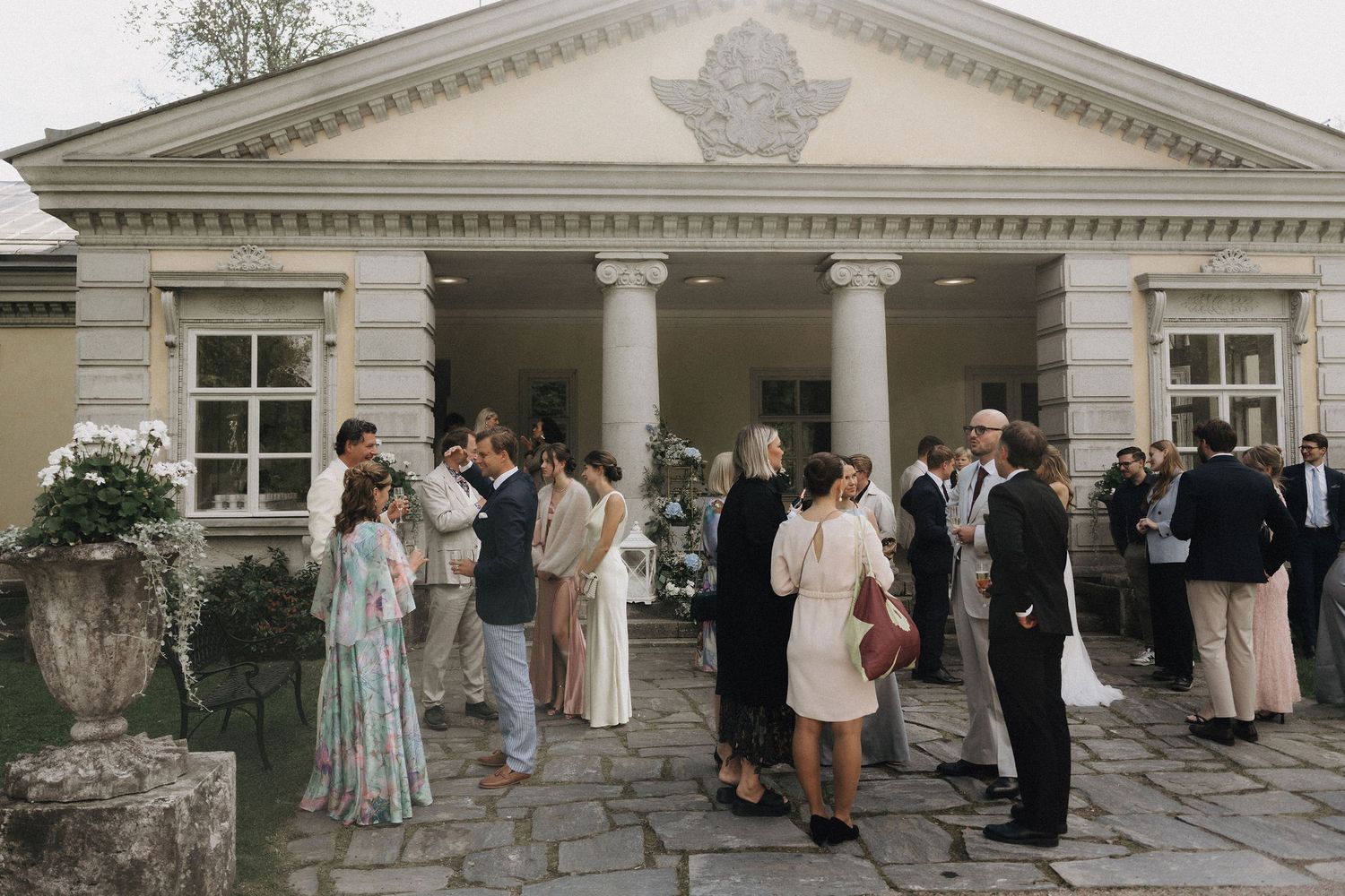 Elegant wedding guests mingling outside a neoclassical venue with white columns and stone urns.